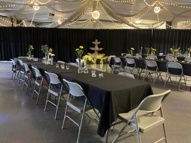 Event space with long tables covered in black tablecloths, decorated with vases of yellow and purple flowers, set with glasses, under a decorated ceiling with string lights and hanging ornaments.
