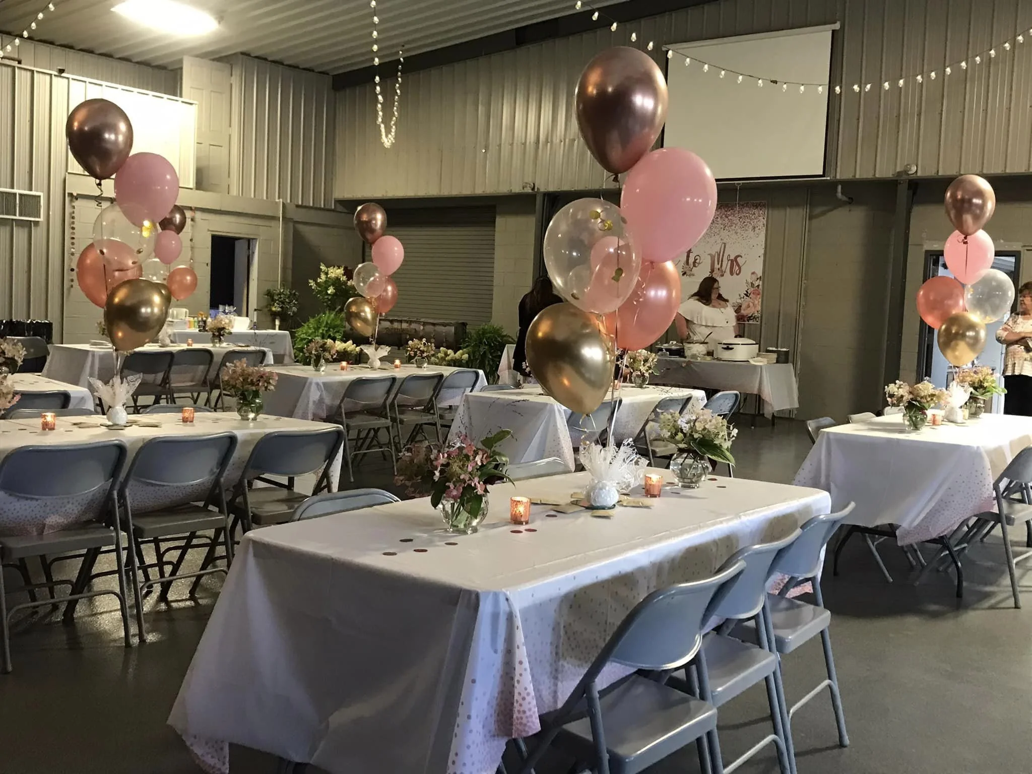 Wedding or celebration reception area decorated with pink, gold, and clear balloons, floral arrangements on tables, and soft lighting in a rustic indoor setting.