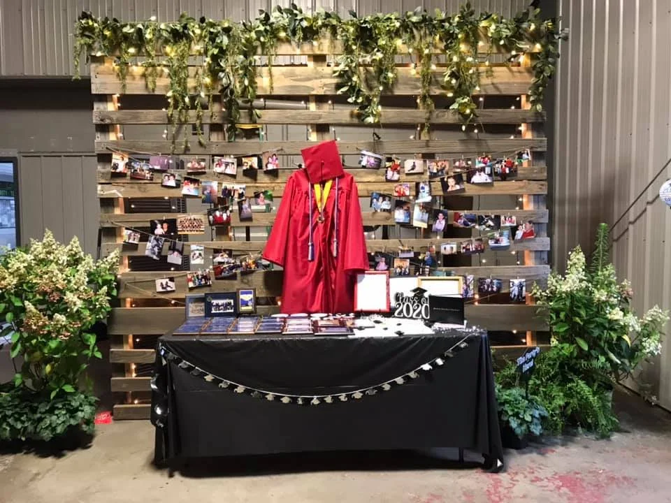 A graduation display with a red graduation gown and cap, medals, and framed photos, set against a wooden pallet backdrop decorated with greenery and string lights, with potted plants on either side.