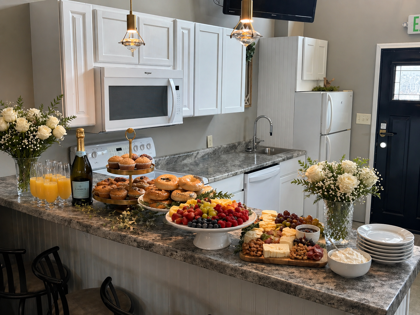 A kitchen decorated for a brunch or breakfast gathering with a marble countertop filled with food, flowers, and drinks. There are fresh fruit, cheese, pastries, juice, and a bottle of champagne. White cabinets, a microwave, and a refrigerator are in the background.