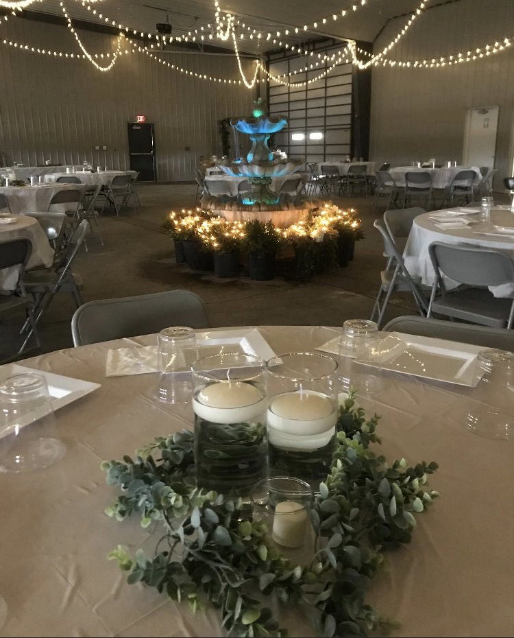 Round banquet table decorated with a candle centerpiece surrounded by greenery, set with glasses and napkins. In the background, a fountain adorned with string lights and greenery, within a large industrial-style event space with additional tables an