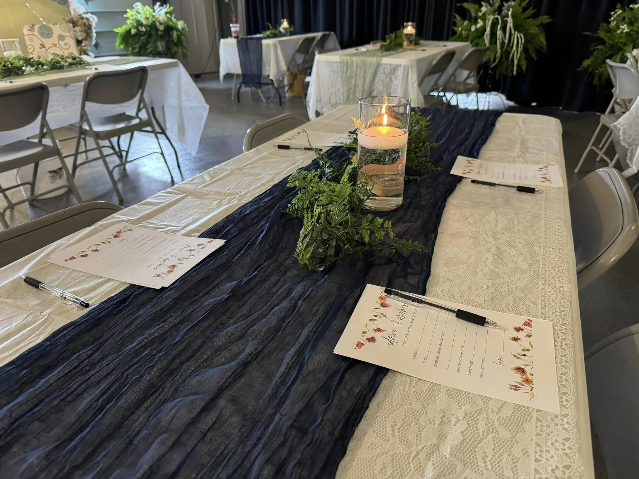 Table decorated with a navy blue runner, greenery, a lit candle in a glass holder, and printed menus or programs.