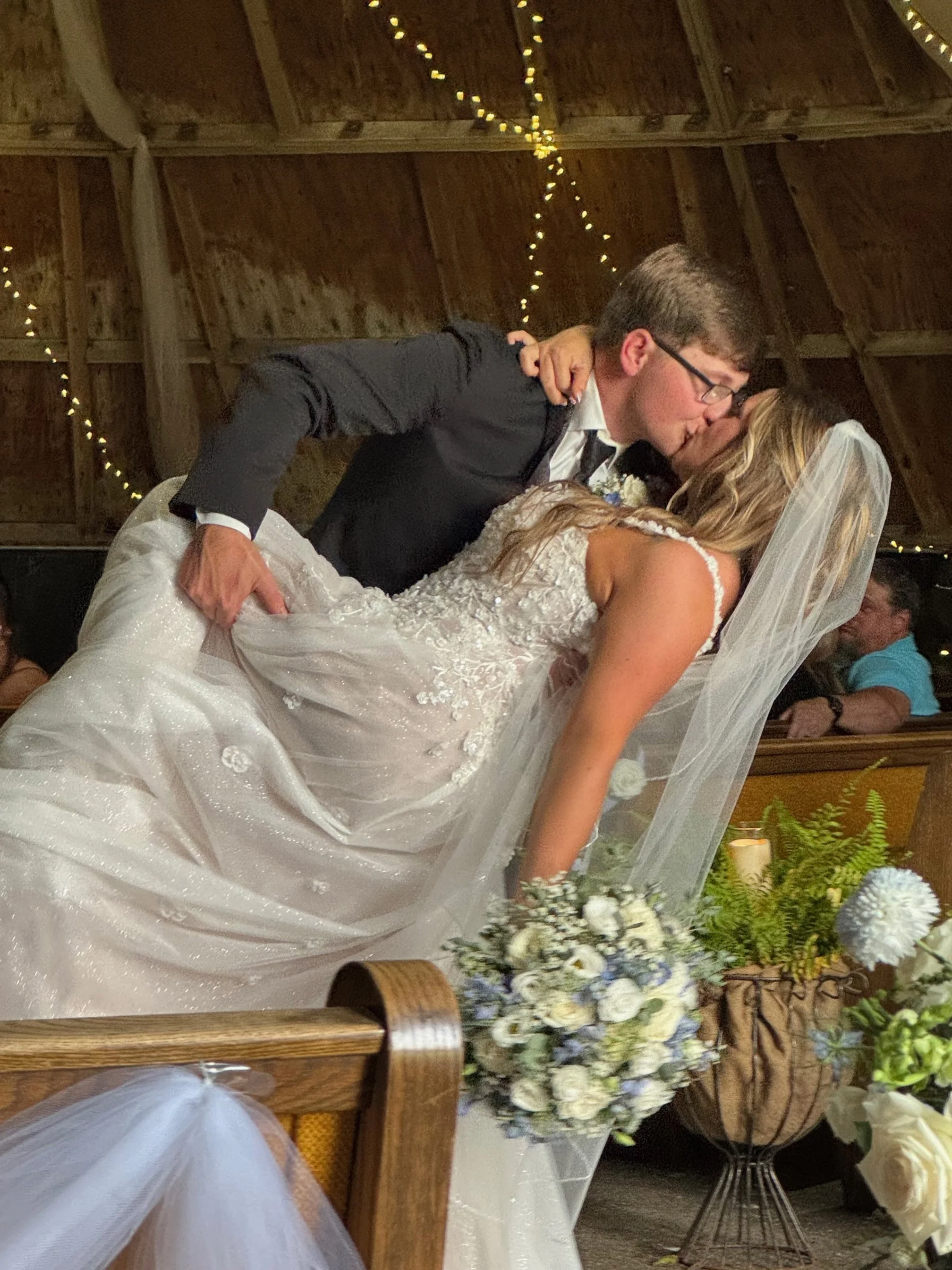 A bride and groom sharing a kiss in a wedding ceremony, with the groom dipping the bride backwards. The groom is wearing glasses and a black suit, the bride is in a lace wedding dress with a veil. There are floral arrangements and candles in the background, with string lights overhead.