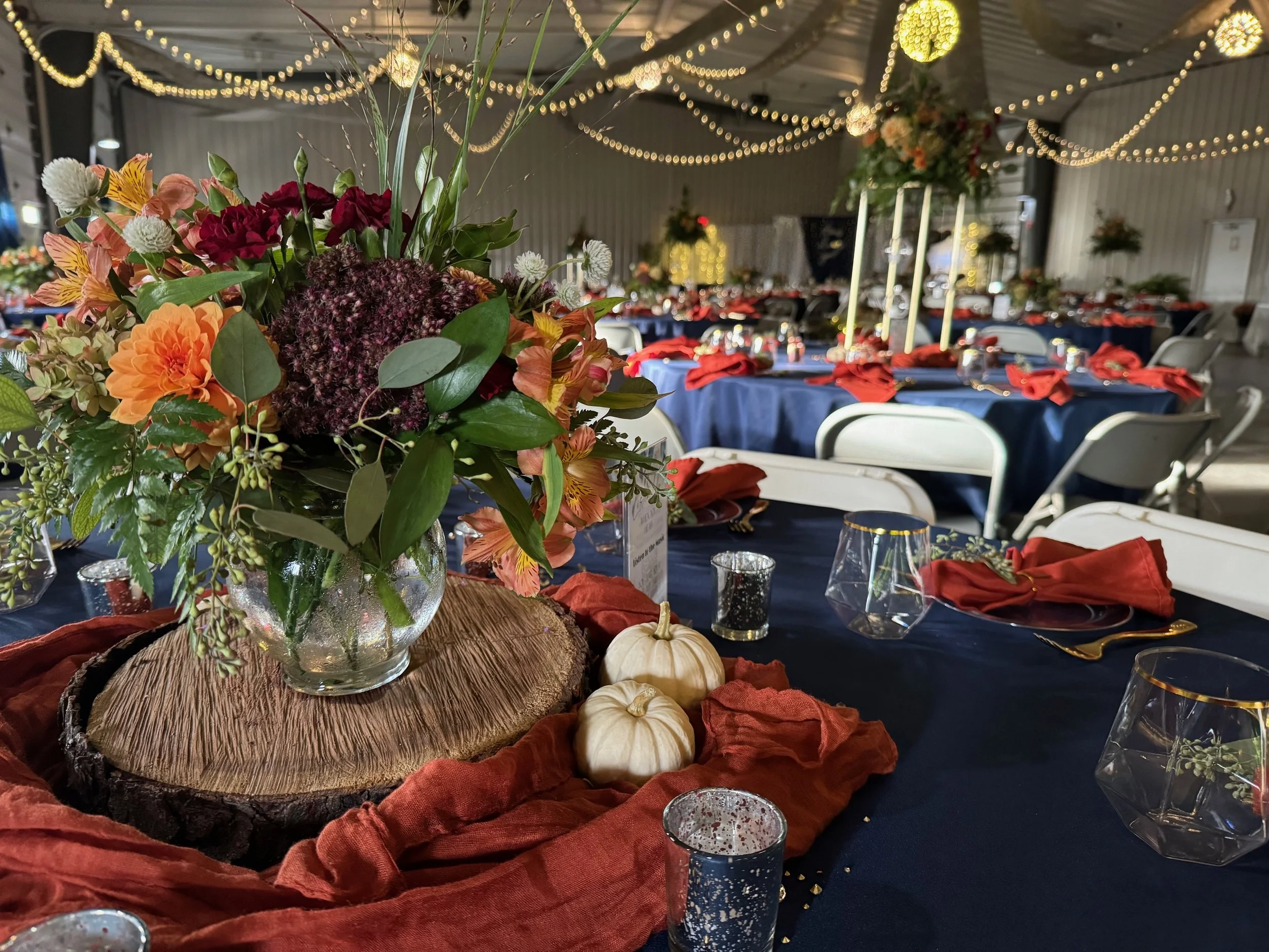 A banquet hall decorated for a celebration with fairy lights hanging from the ceiling. The tables are covered with navy blue tablecloths and red napkins. The centerpiece is a large floral arrangement with various flowers and green foliage, placed on 