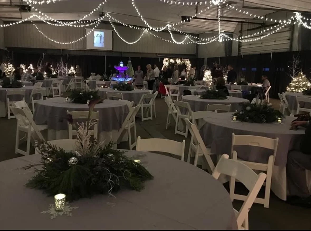 A decorated indoor holiday event space with round tables covered in white cloths, adorned with festive greenery centerpieces, and surrounded by white chairs. The ceiling is decorated with string lights, creating a warm, festive atmosphere. There are 
