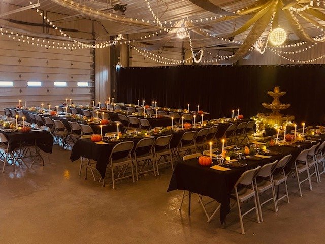 Indoor event space decorated for a fall celebration with long tables covered in black tablecloths, orange pumpkins, candles, and a three-tiered fountain.

String lights and fabric drapes adorn the ceiling, creating a festive atmosphere.