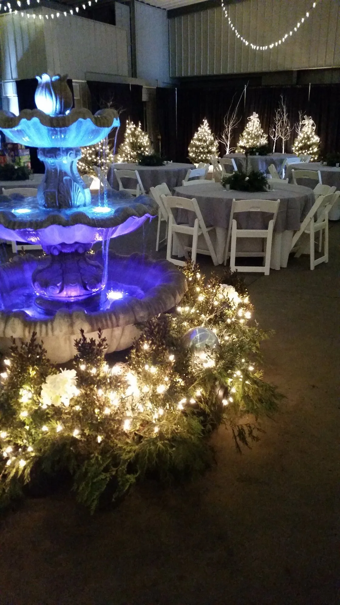 Indoor holiday celebration with illuminated Christmas trees, a multi-tiered fountain with blue and purple lighting, decorated with white flowers and string lights.