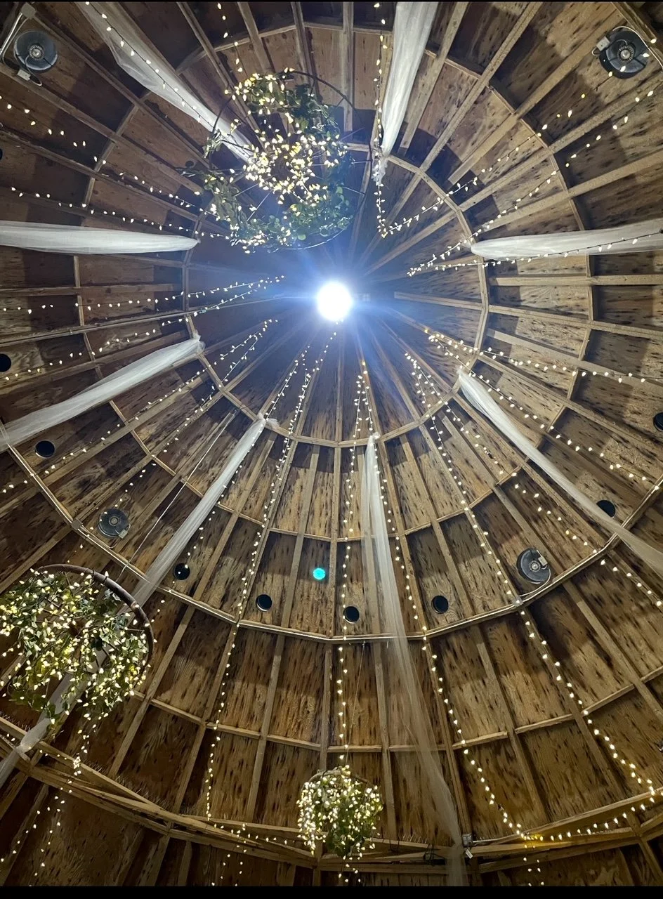 View from the inside of a wooden circular ceiling with hanging string lights and floral arrangements.
