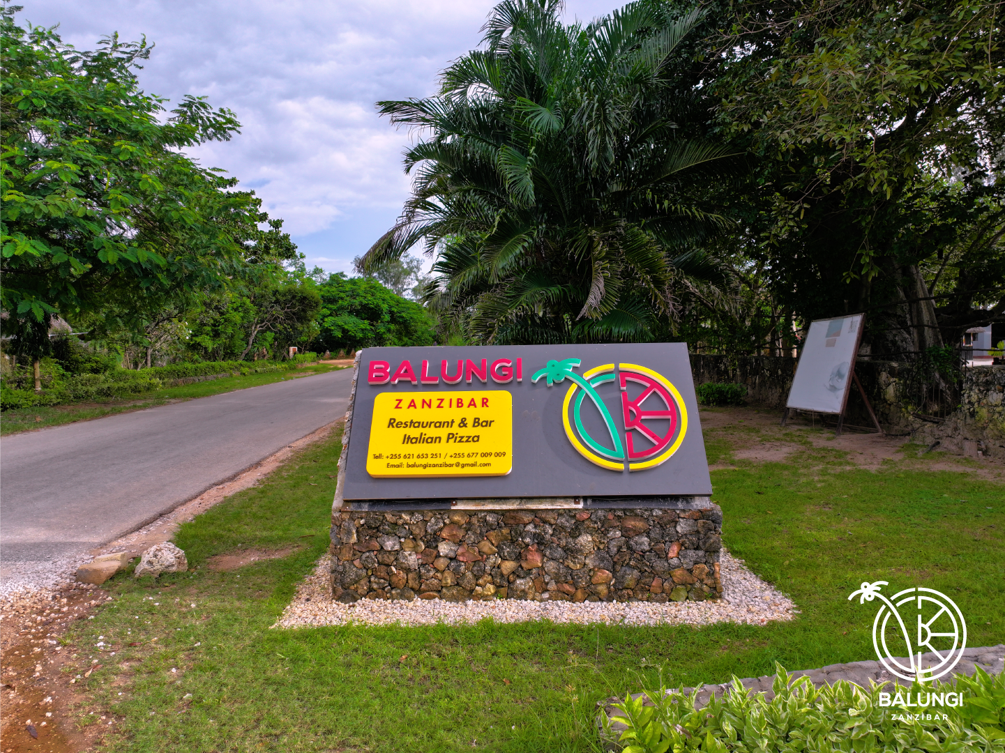 Signboard advertising Balungi Zanzibar restaurant and bar, offering Italian pizza. The sign is mounted on a stone base, with colorful text and graphics, including a stylized lemon and bicycle. The background features trees, greenery, and a cloudy sky