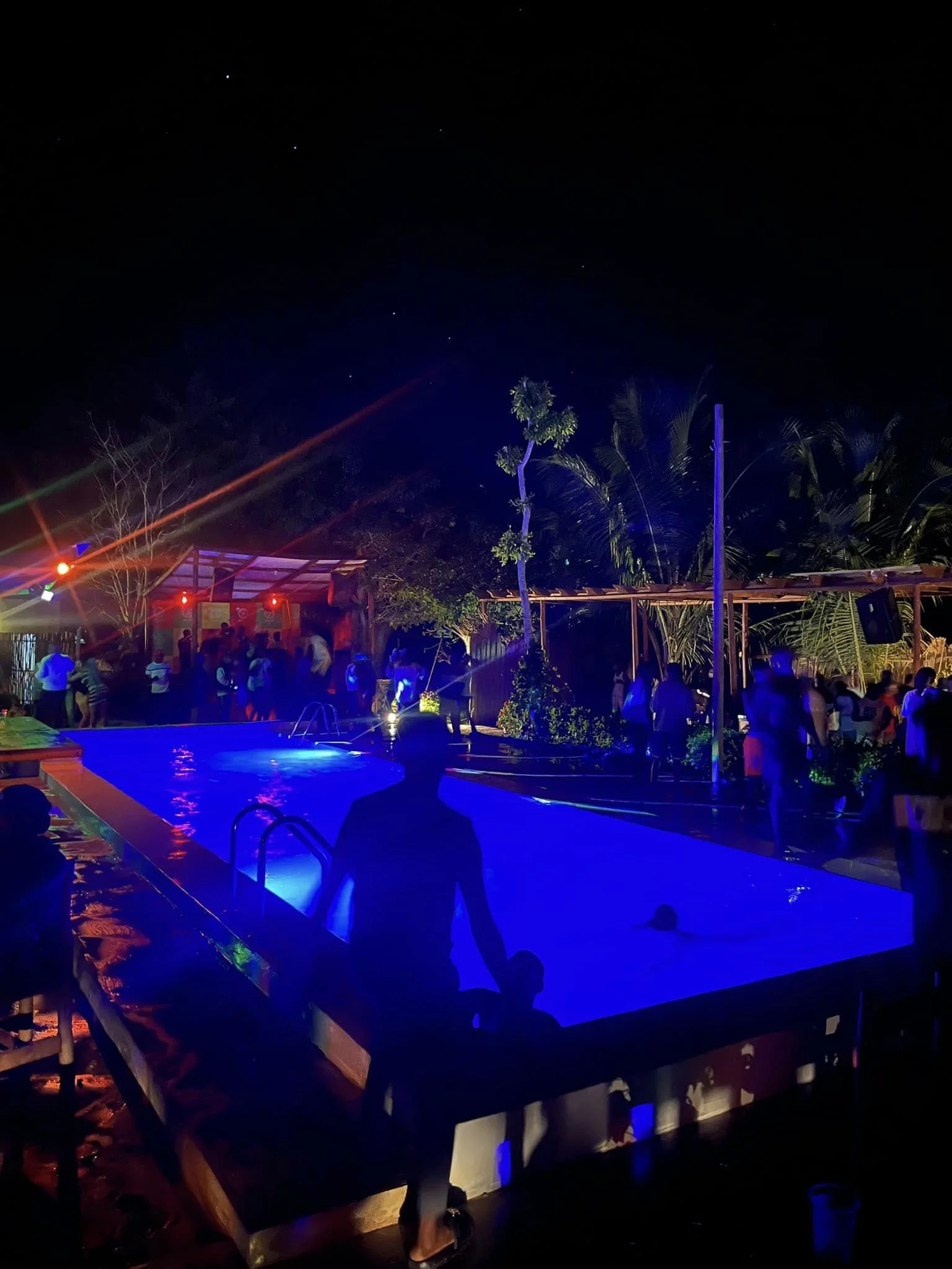 Nighttime scene at a pool party with illuminated blue pool, people socializing and dancing under string lights, surrounded by tropical trees.