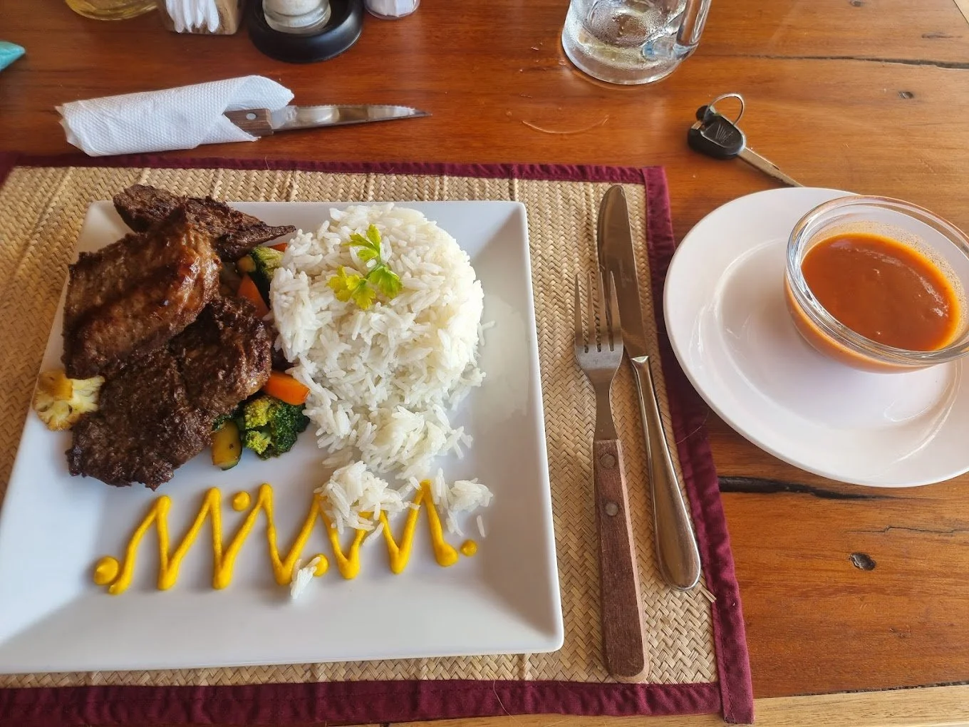 Plate of rice with meat and vegetables, mustard decoration, bowl of soup, fork, knife, and keys on a wooden table.
