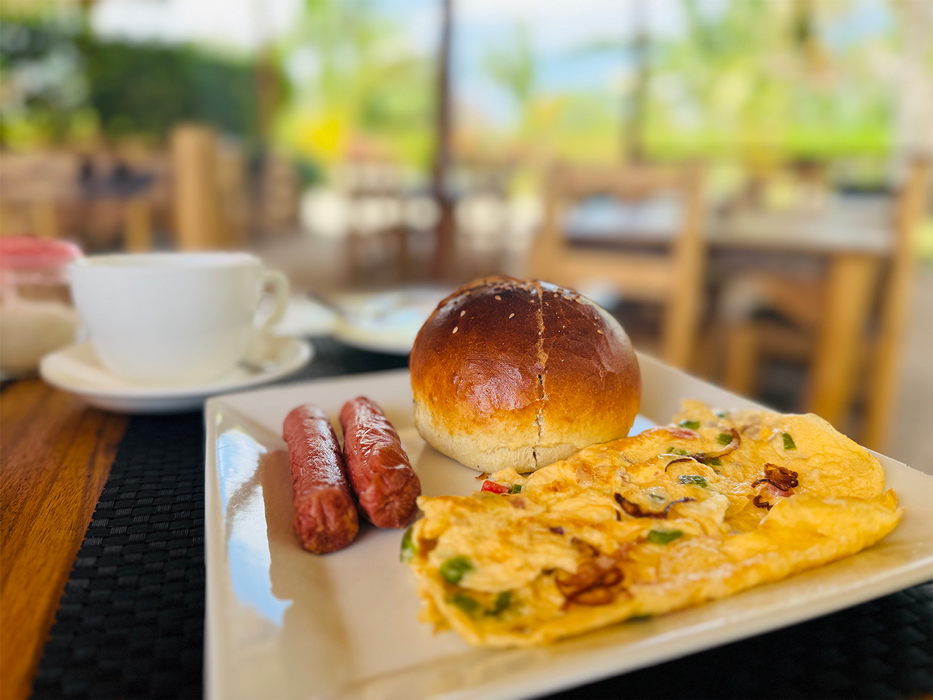 Breakfast plate with two sausages, an egg omelet with vegetables, and a bread roll, on a white tray at a restaurant table.