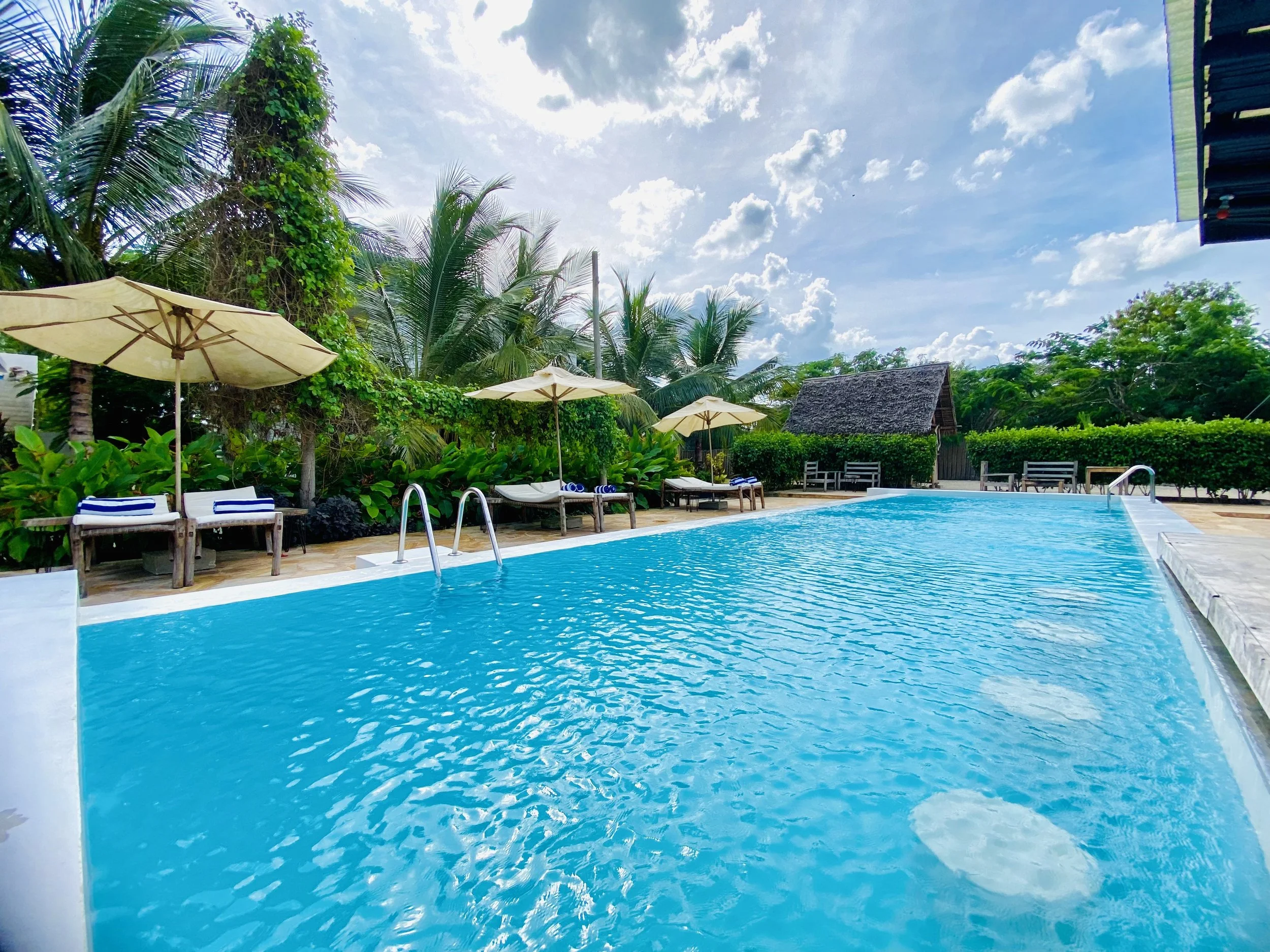 Swimming pool with lounge chairs in a lush tropical setting under a partly cloudy sky.