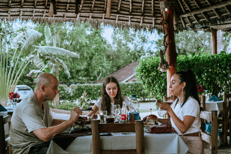 Three people dining outdoors at a restaurant with tropical greenery and thatched roof overhead.