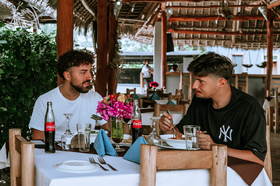 Two men having a meal at a restaurant, one with curly hair and beard, the other with straight hair, sitting at a table with flowers, drinks, and plates of food, in a tropical setting with a thatched roof.