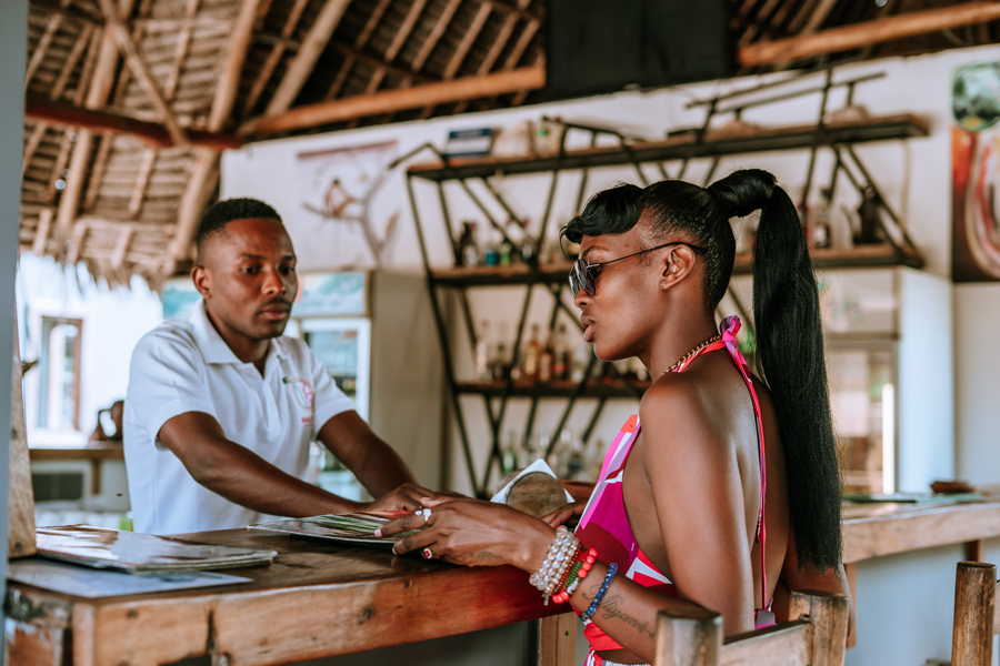 A woman at a bar counter handing money to a bartender in a tropical bar with thatched roof and wooden shelves.