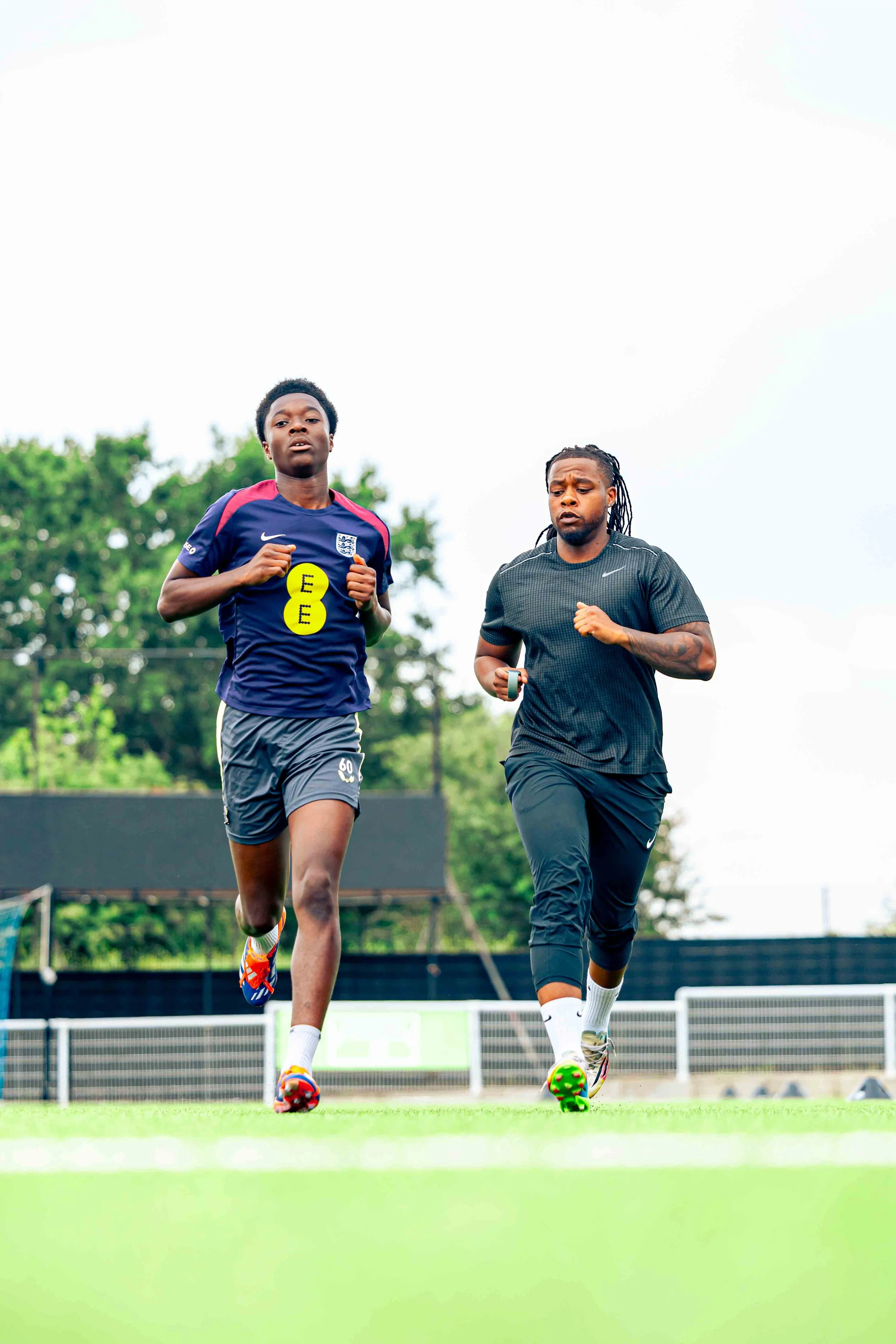 Two men running on a grass field during daytime, one in a blue sports jersey and the other in black athletic wear.