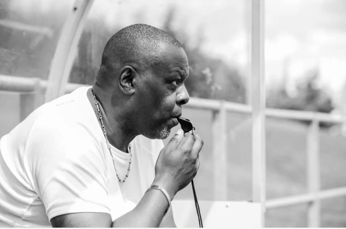A man in a white shirt using a whistle during a sports event, sitting on a bench near a transparent barrier.