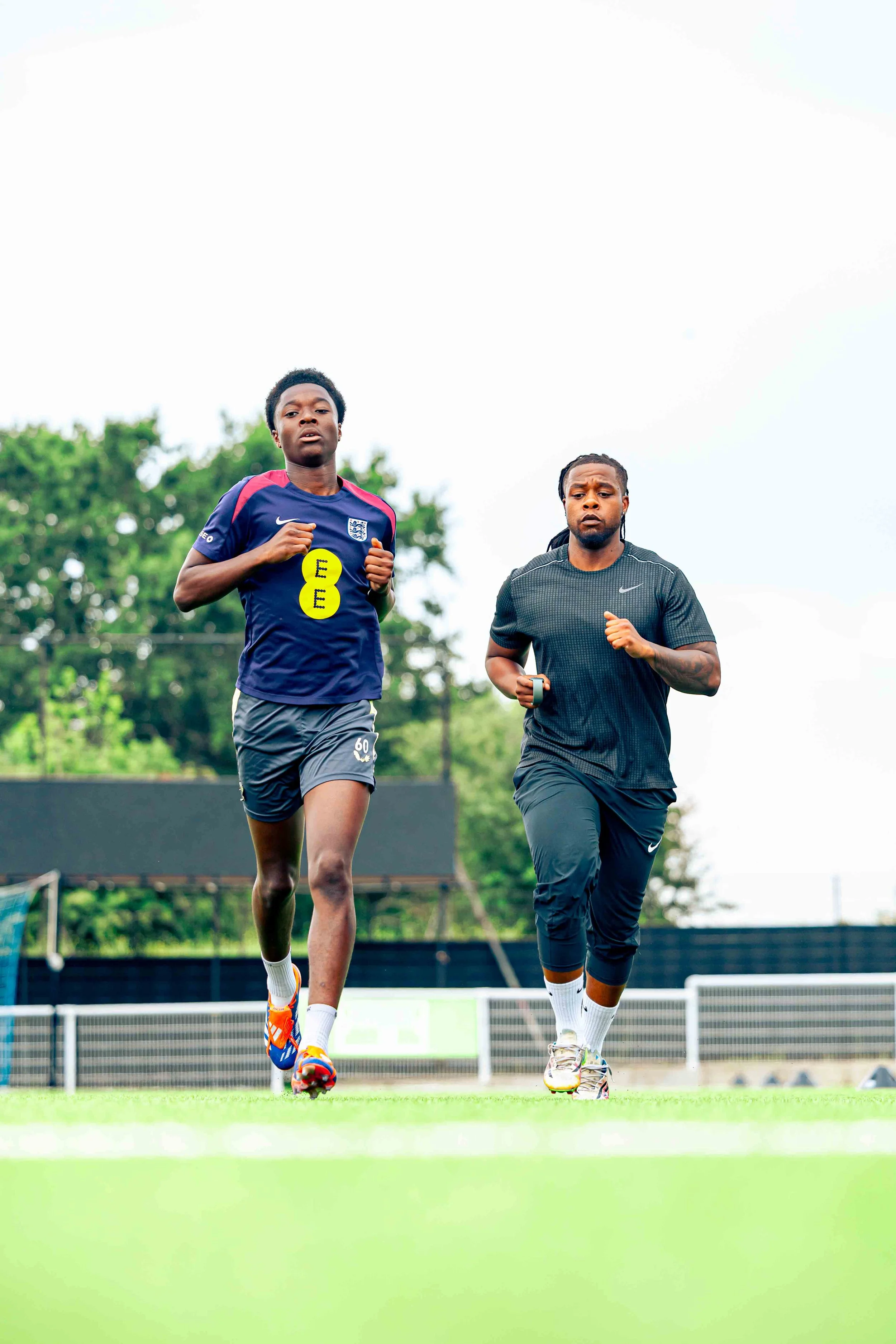 Two men jogging on a sports field during daytime, with trees and a black fence in the background.