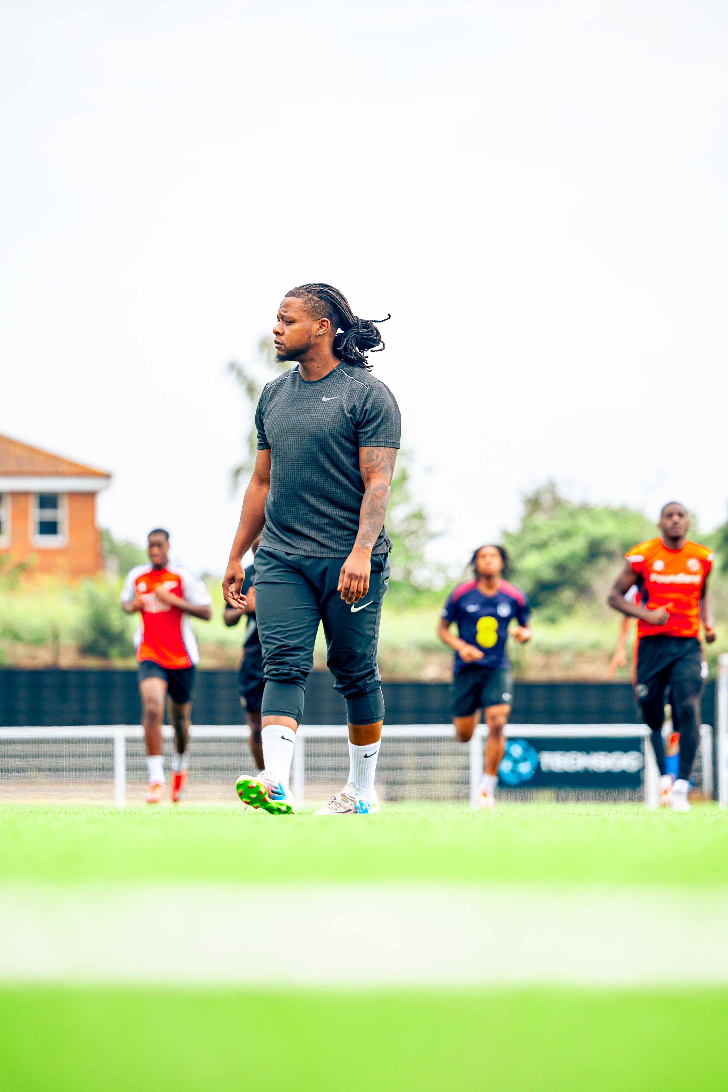 A man in athletic gear, standing on a soccer field during a training session, with other players in the background.