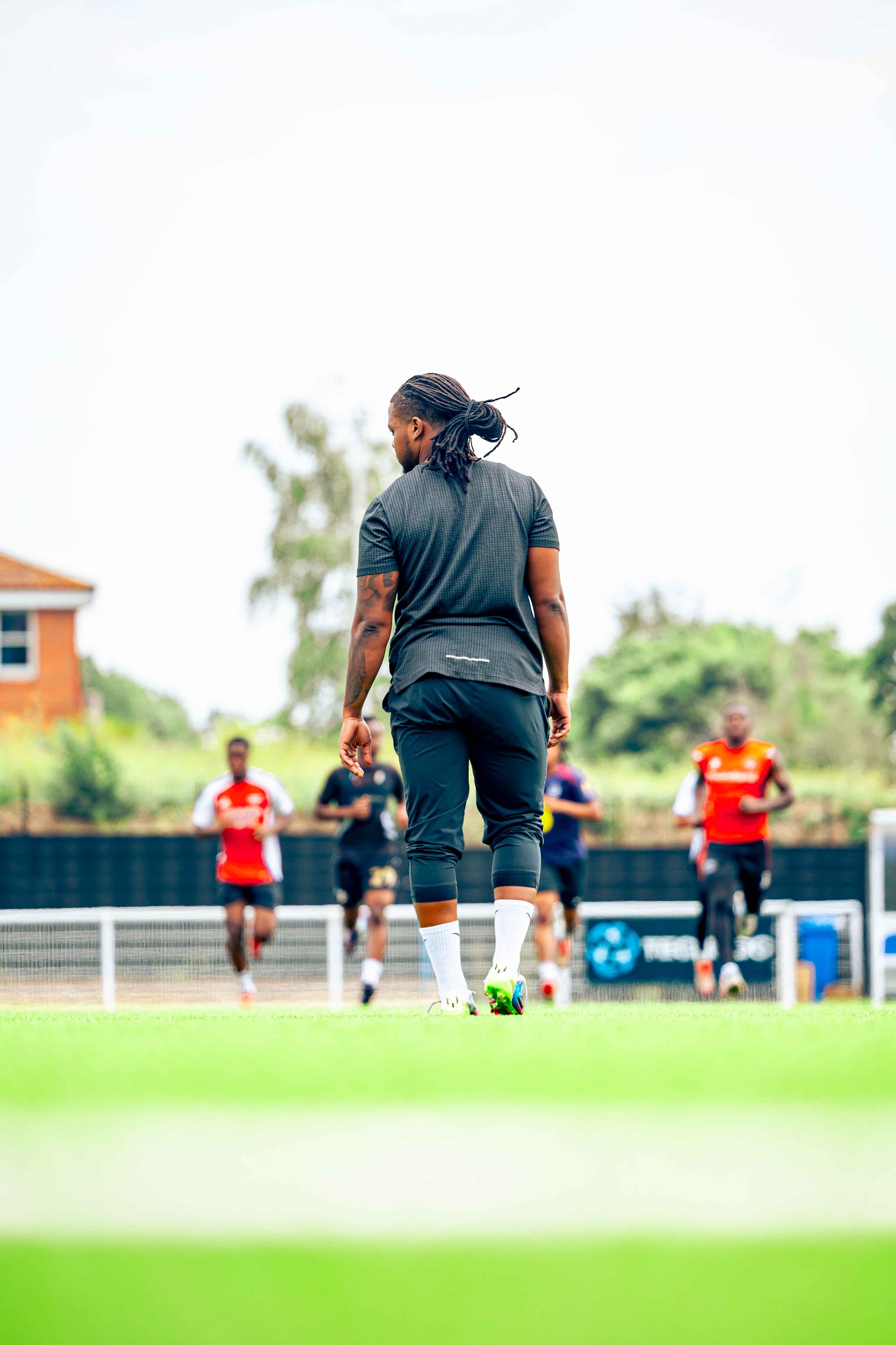 A soccer coach or trainer standing on a soccer field with players in the background during practice or training session.