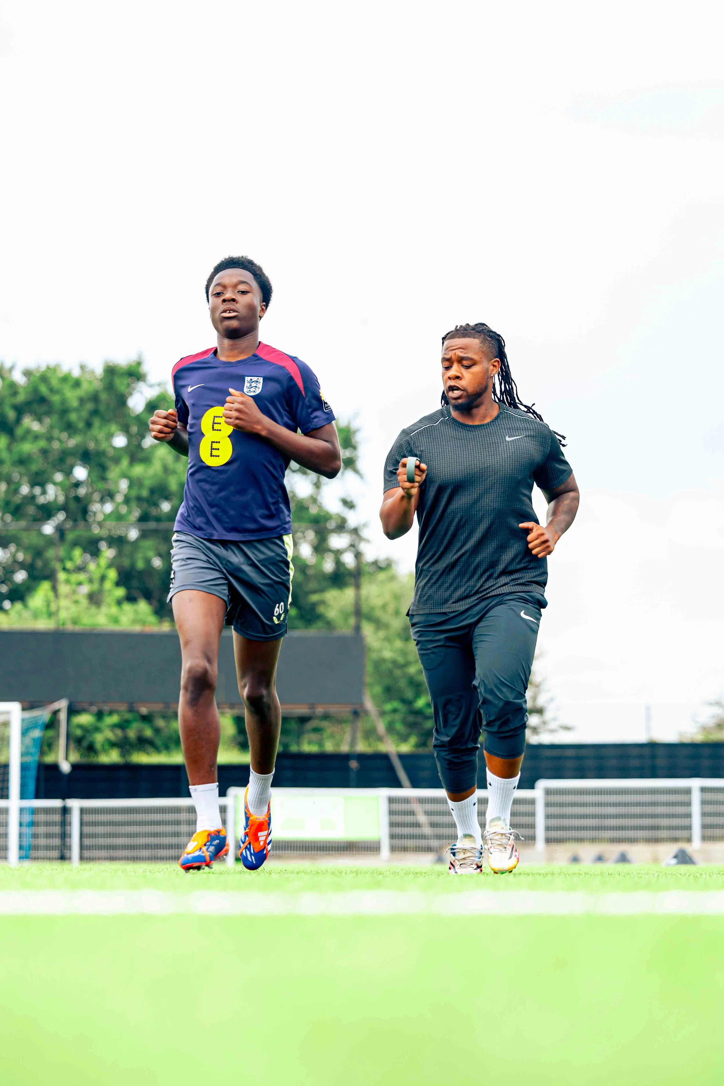 Two men jogging on a football field, with one wearing a blue sports jersey and the other in a black sports shirt.