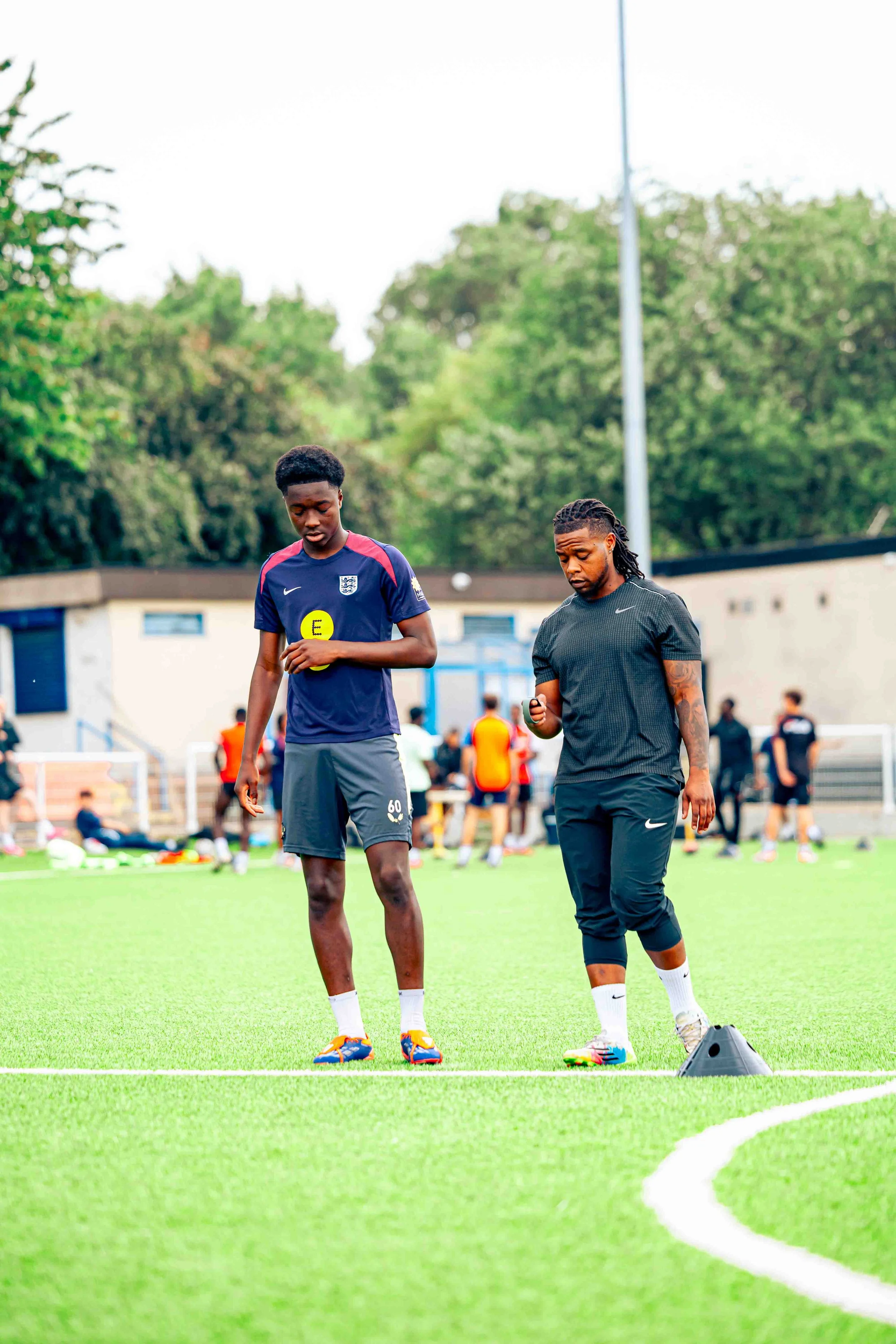 Two soccer players standing on a field during training, one holding a yellow ball marker, with other players and coaches in the background.
