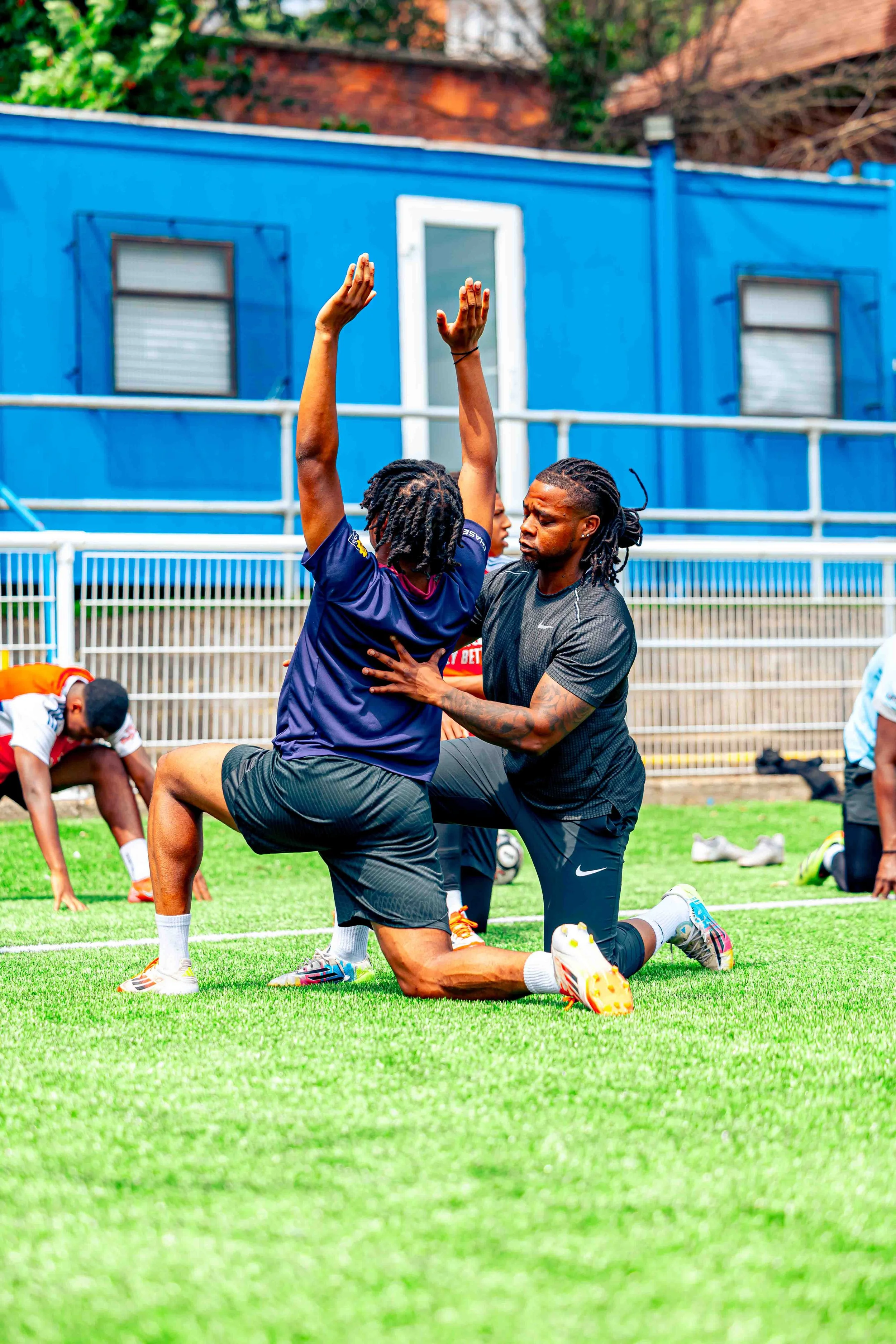 Two men practicing football drills on a field, with one man helping the other stretch; other players are in the background