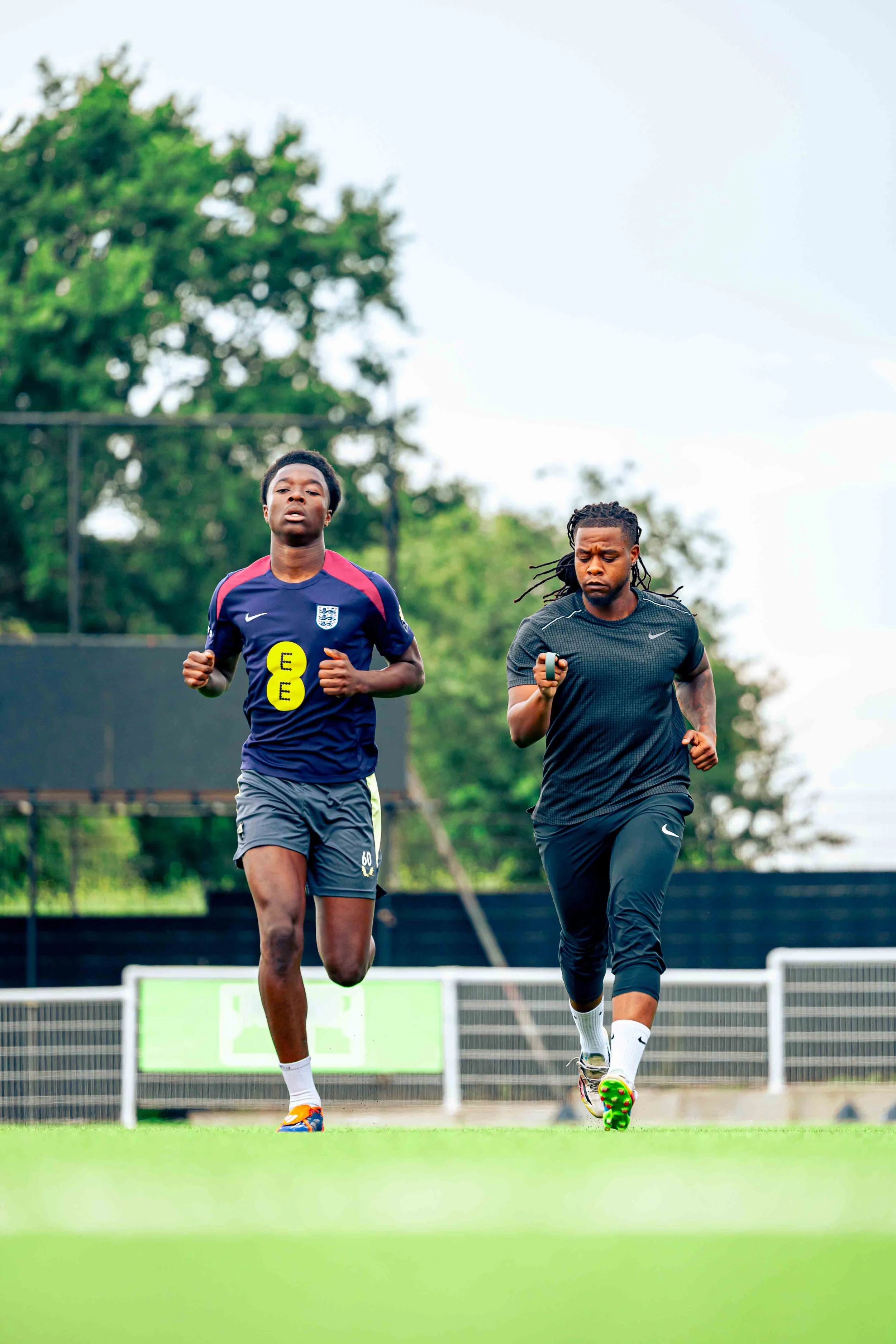 Two young men jogging on a soccer field, with trees and a scoreboard in the background.