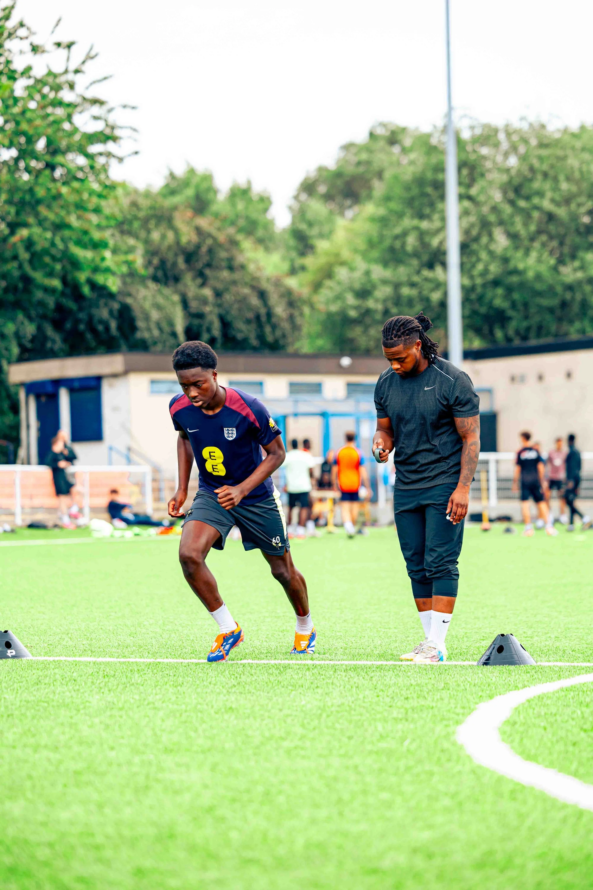 Two young men performing agility drills on a green football field with cones, while other people gather in the background.