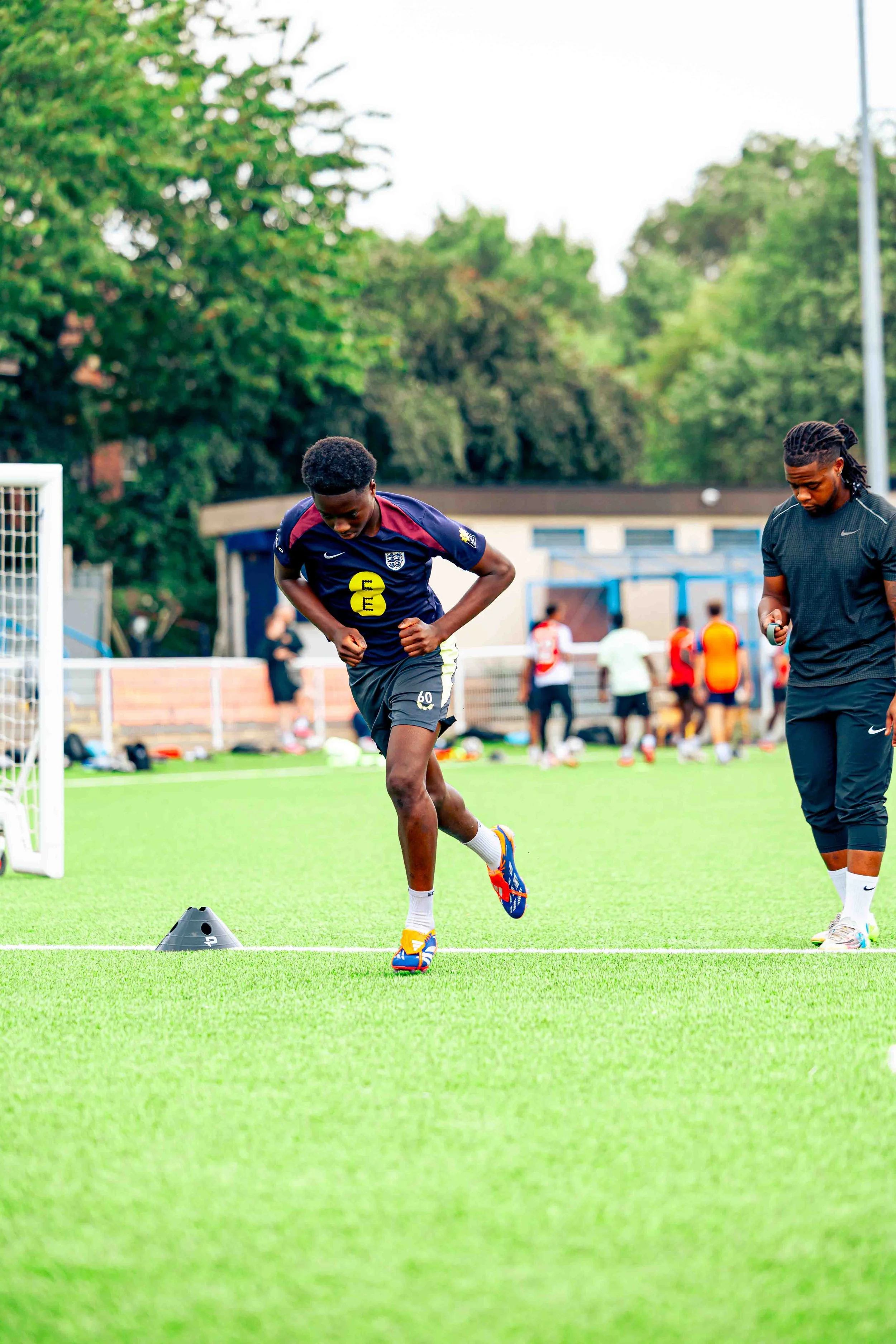 A young male soccer player practicing on a field, running between cones, with a coach or trainer nearby checking their phone, and other players in the background.