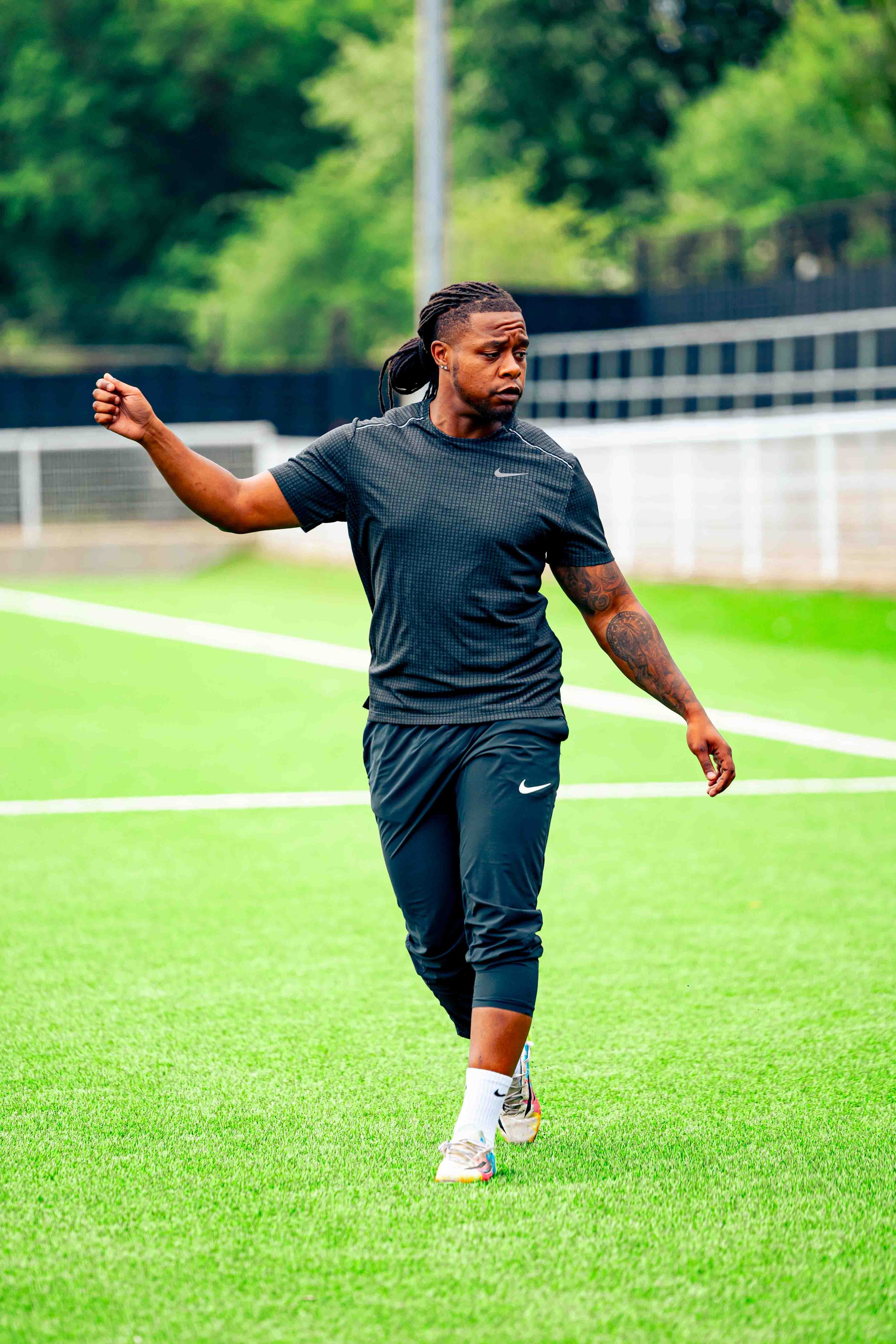 A man with dreadlocks and tattoos on his left arm is standing on a football field with green grass. He is dressed in black athletic clothing with Nike logo and white Nike shoes, appearing to be warming up or stretching.