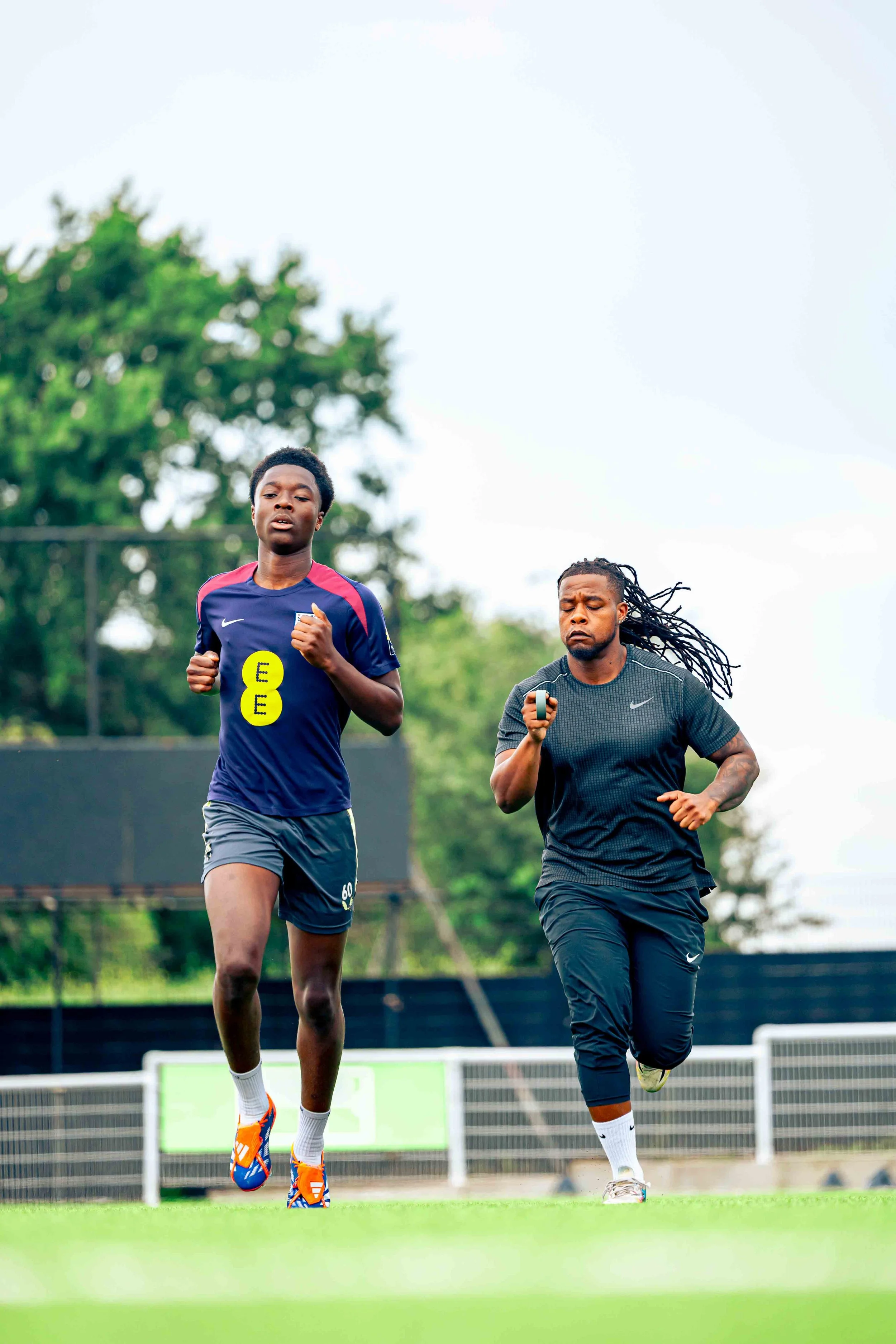 Two athletes, a young man and a woman, running on a sports field during daytime with trees in the background.