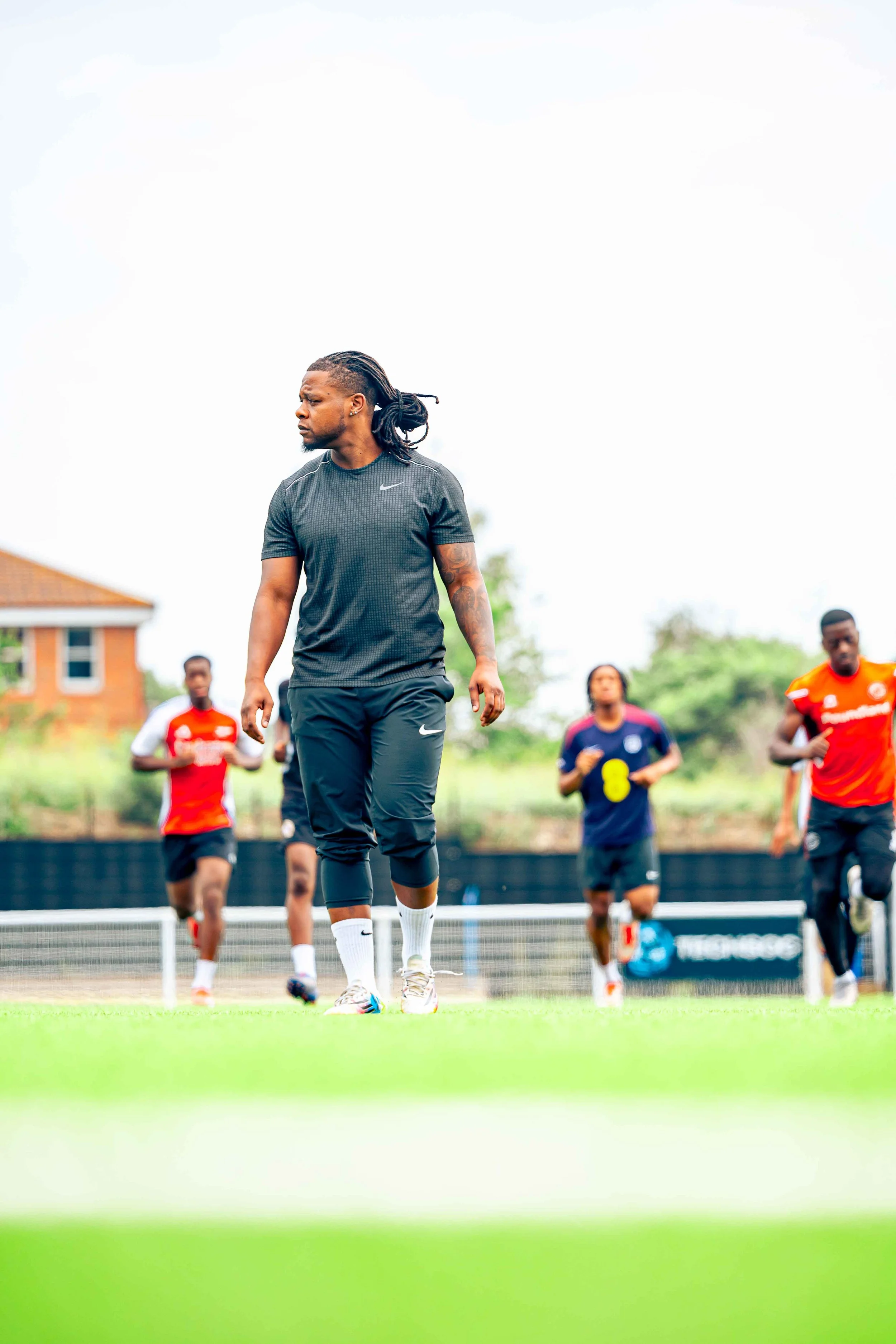 A coach or player in athletic gear walking on a soccer field, with four other players running in the background during a training session.