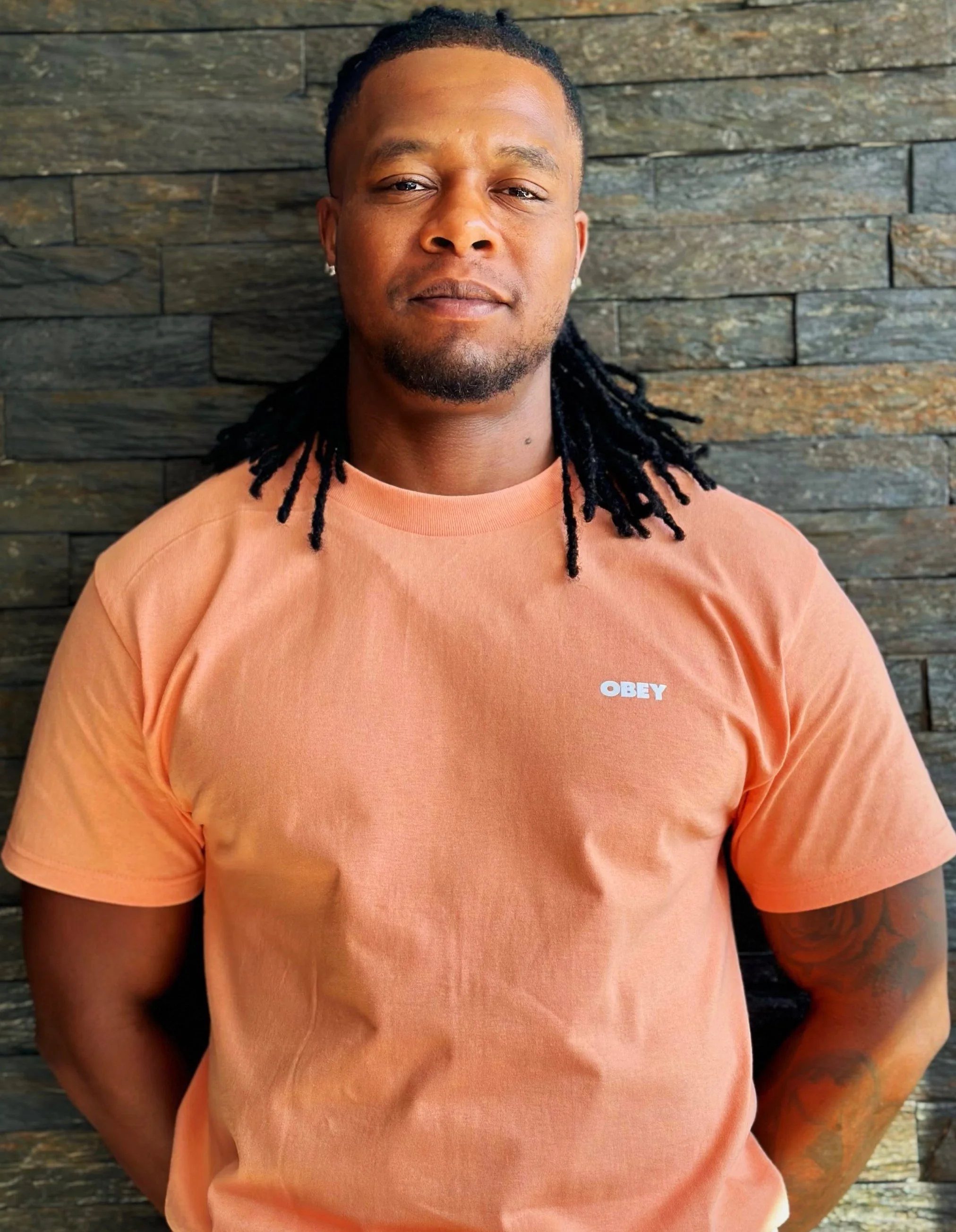 A man with dreadlocks wearing an orange t-shirt with the word "OBEY" on it, standing against a stone wall.