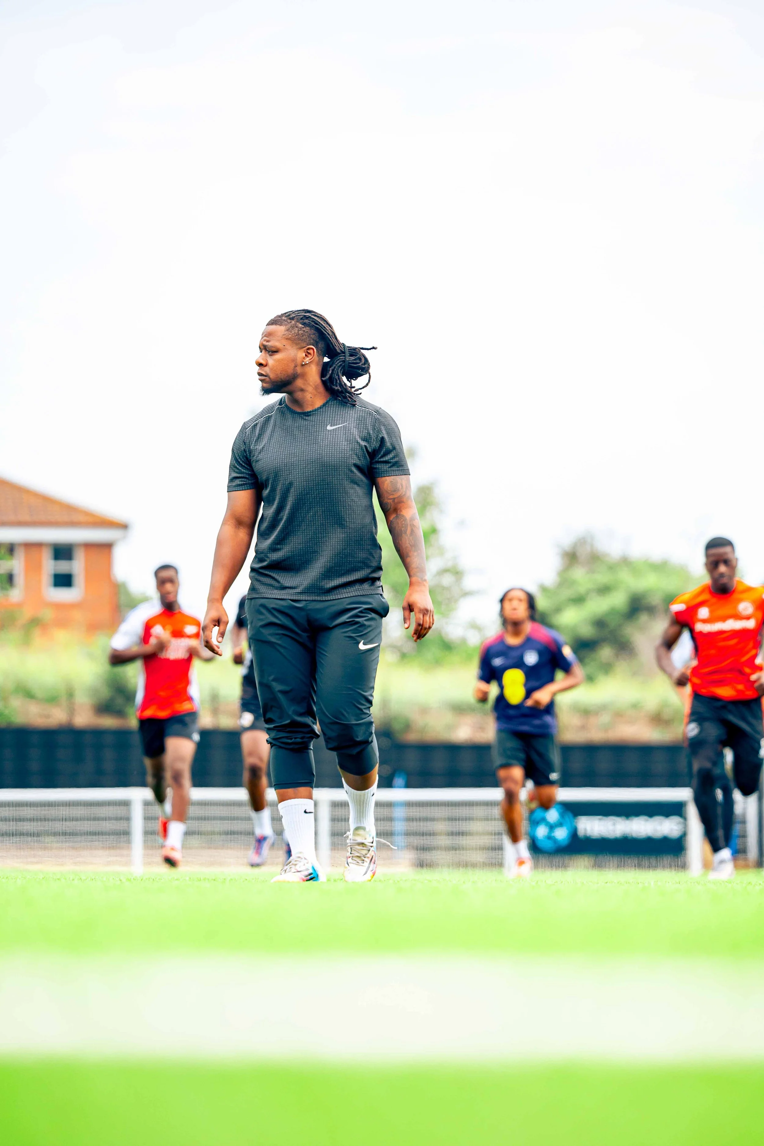 A man in athletic wear standing on a soccer field with a group of other players running behind him during practice or a game.