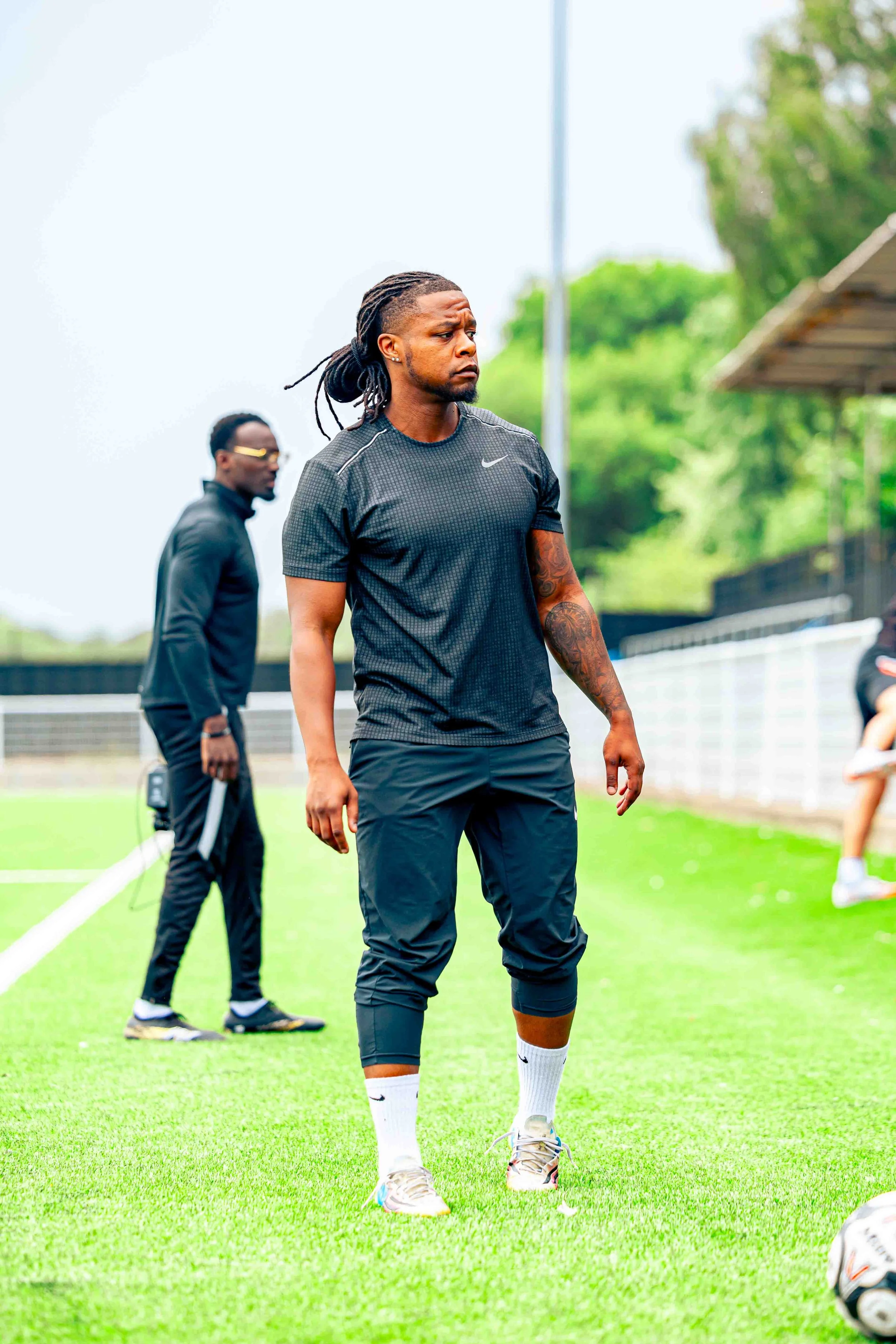 A soccer player with long dreadlocks wearing a black athletic shirt, black pants, and white socks standing on a green soccer field, looking to the side. Another person in the background is also on the field.