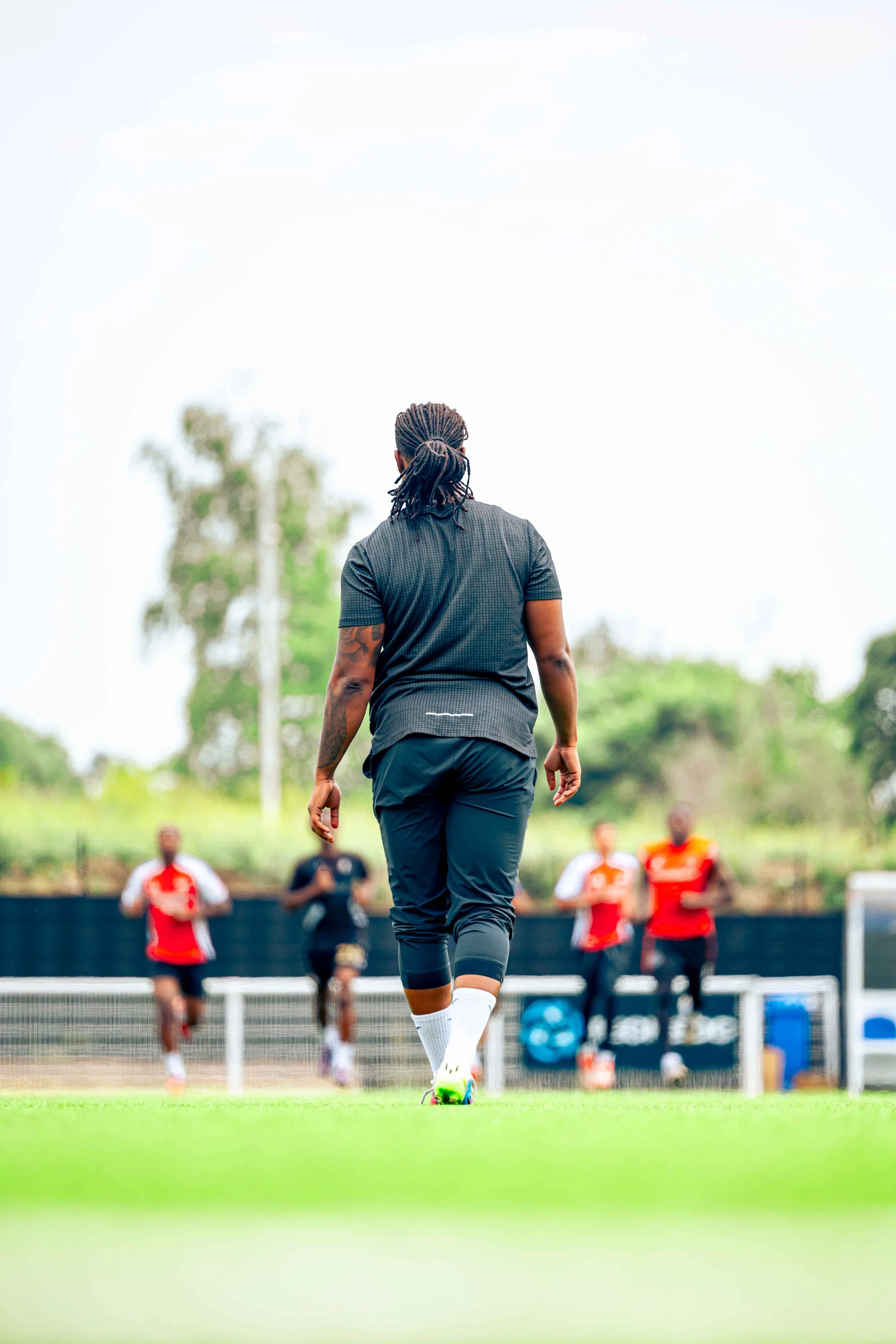 A person with dreadlocks walking on a sports field with four other players running in the background.
