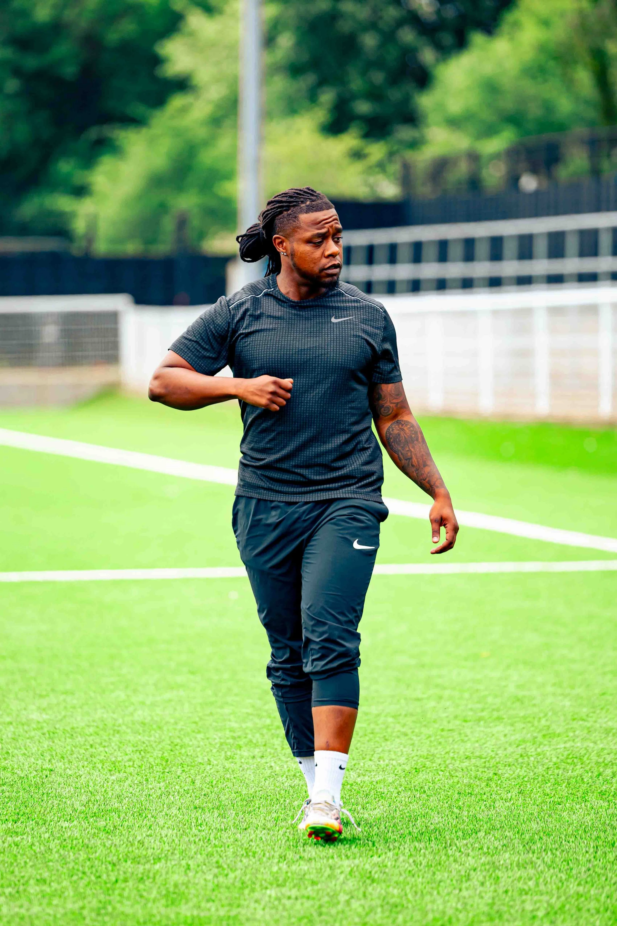 A man in sports attire is running or jogging on a grassy field during daytime, with trees and a stadium or sports facility in the background.
