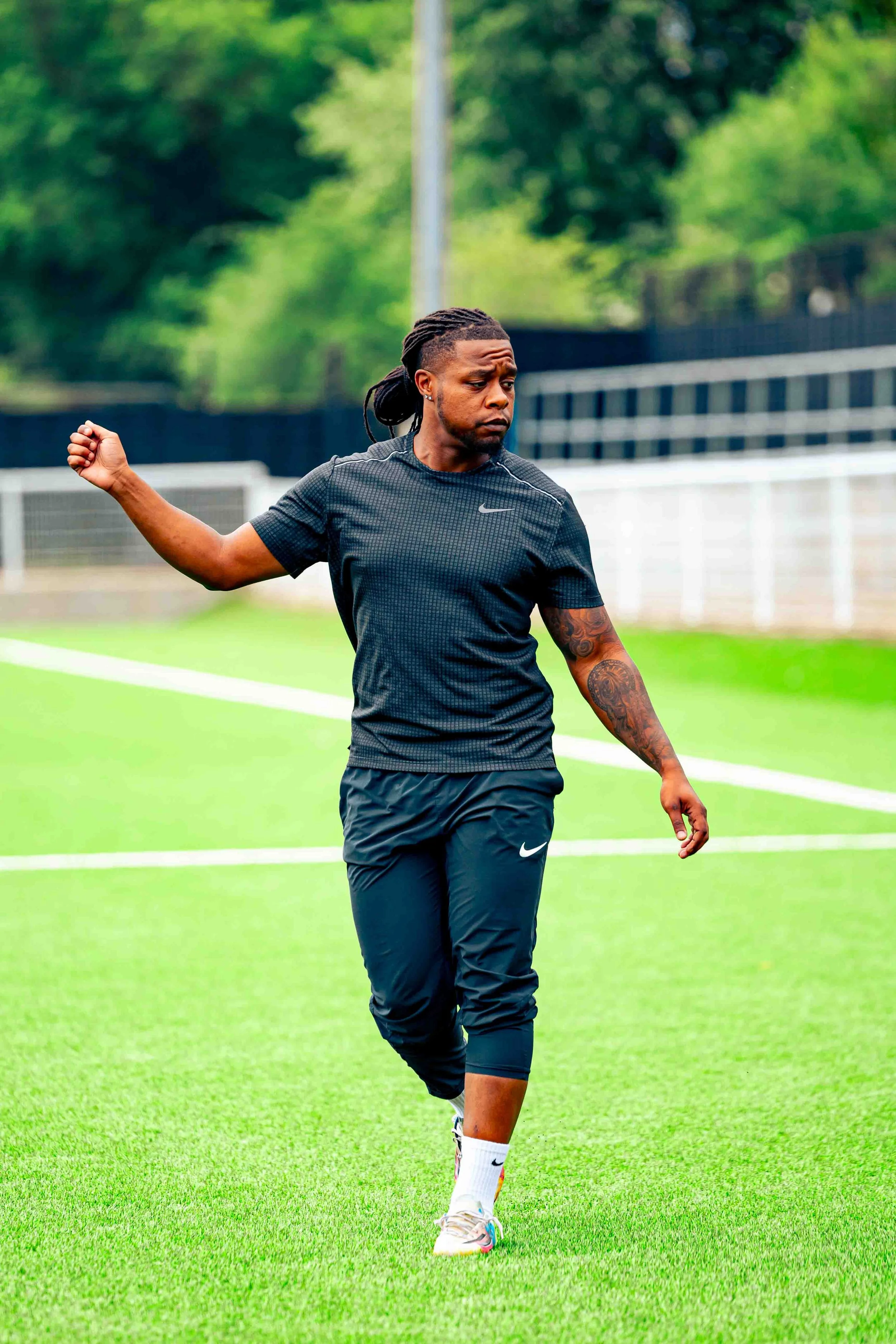 A man with dreadlocks, athletic build, in a black Nike sports outfit, walking on a green soccer field with trees and a fence in the background.