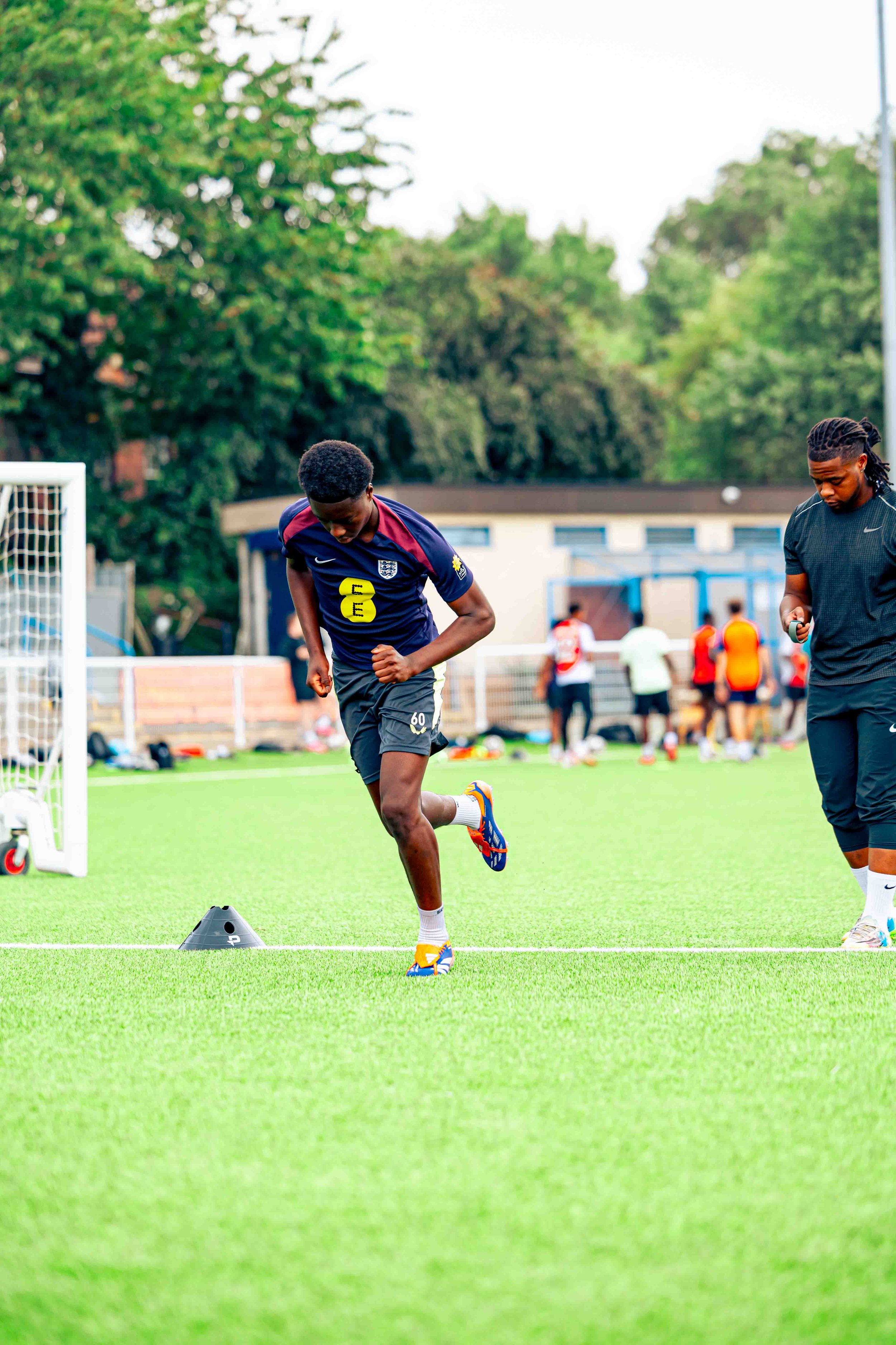 A young male athlete, wearing a navy blue sports jersey with yellow numbers and patches, is running on a green soccer field during a training session. There are cones on the ground and other players and a coach in the background.