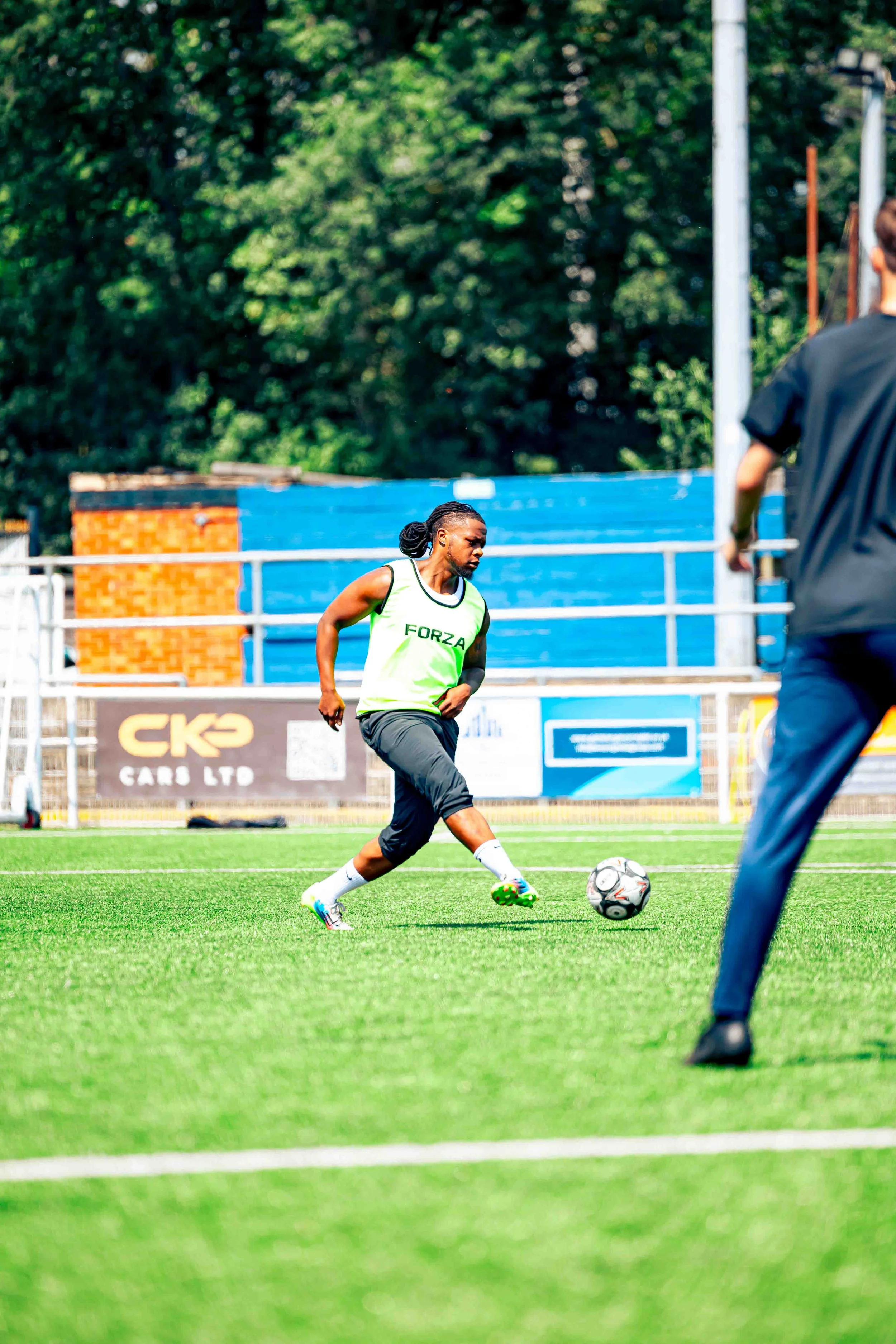 A female soccer player in a green and white jersey, black shorts, and white socks, about to kick a soccer ball on a green field, with another person partially visible in the foreground, and trees and advertisements in the background.