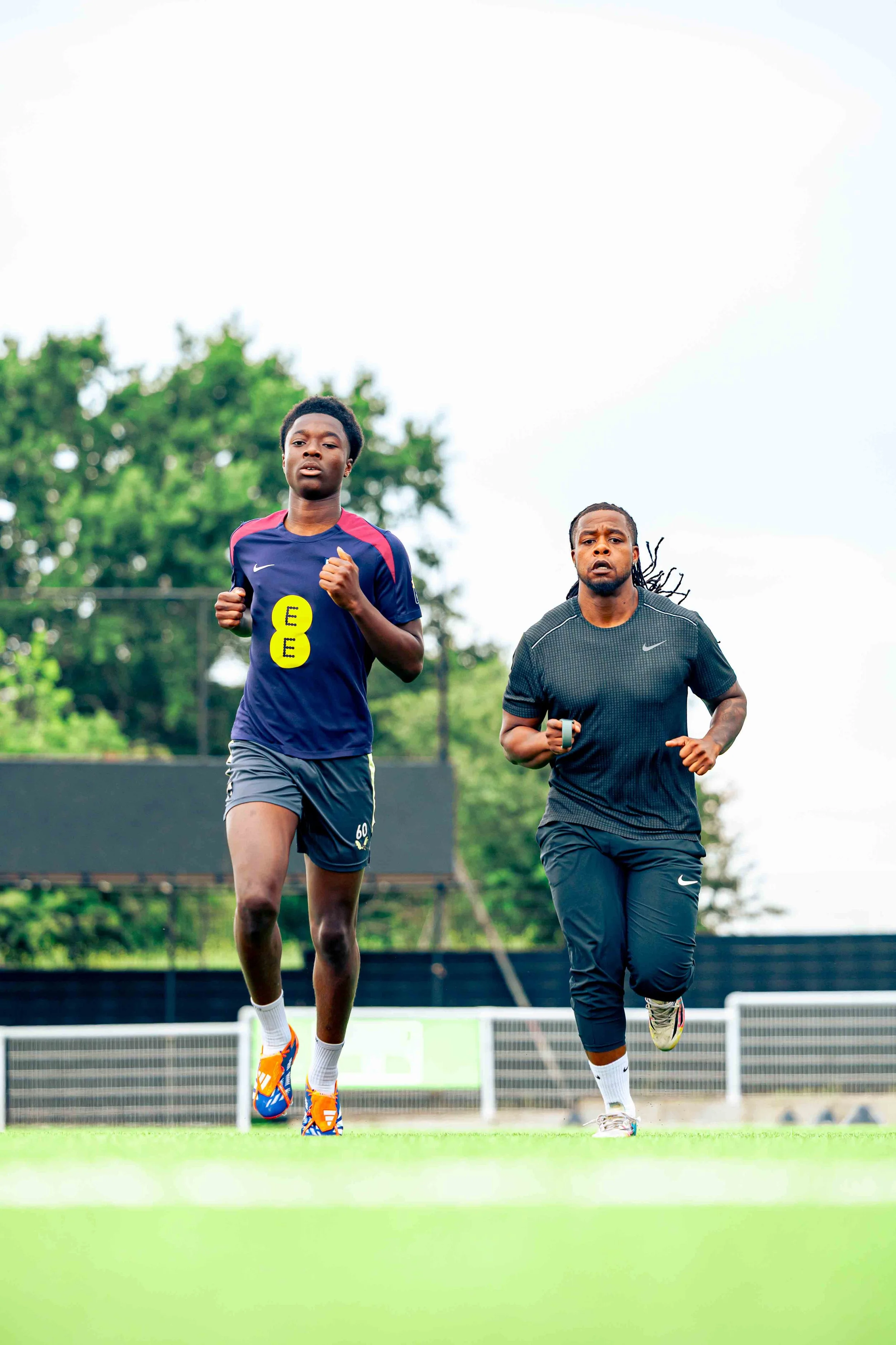Two men jogging on a running track outdoors, with green trees in the background, wearing athletic clothes.