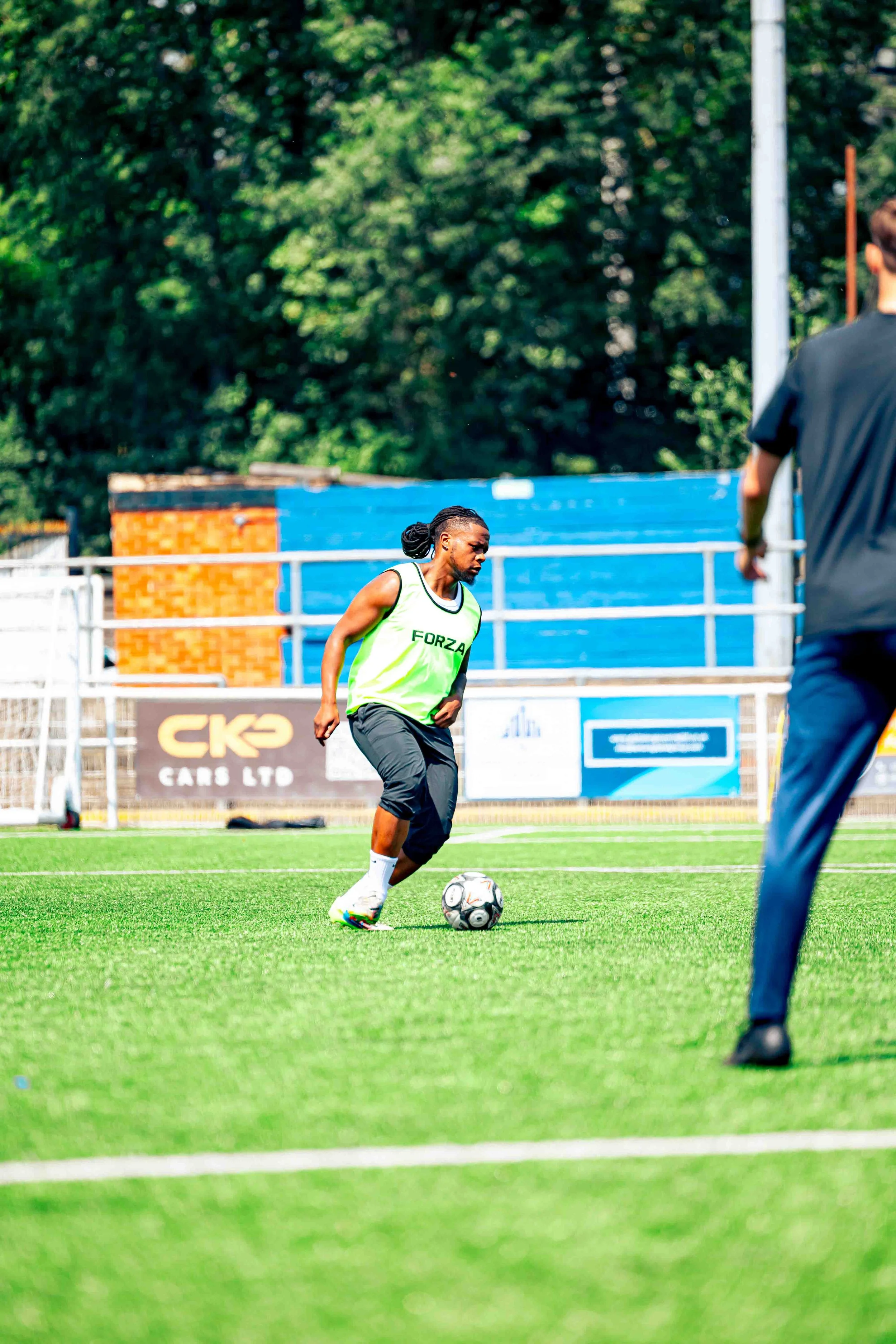 A woman playing soccer on a field, wearing a green vest labeled 'FORZA', dribbling a soccer ball, with a man in dark clothing in the foreground and trees in the background.