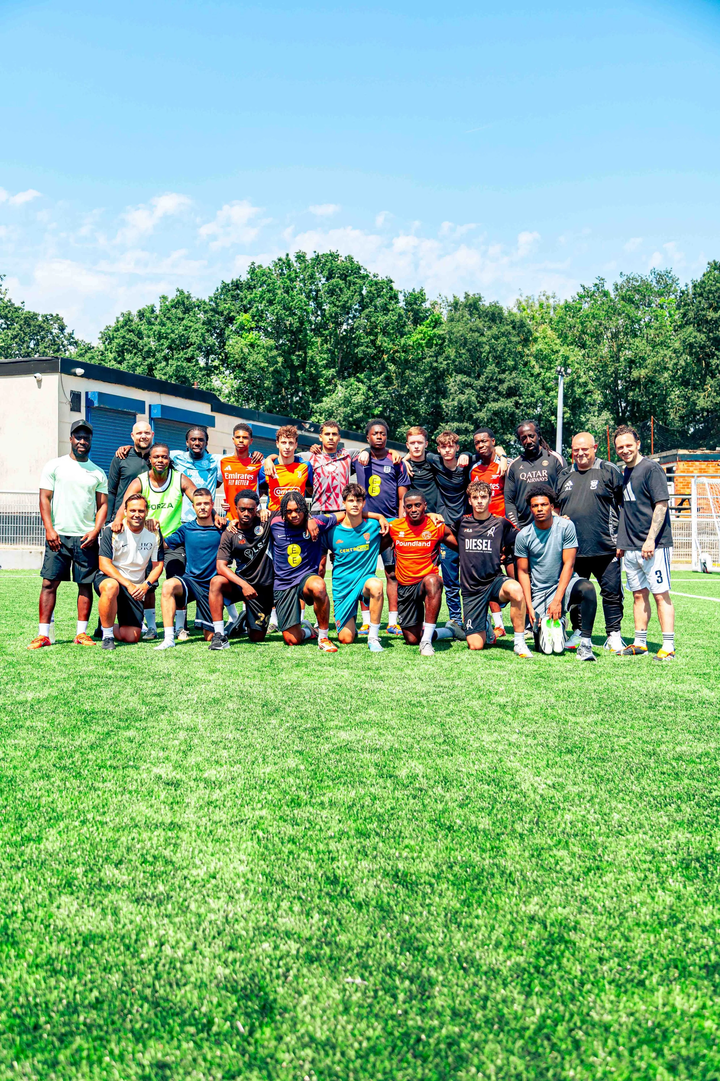 Group of people, including young athletes and coaches, posing for a photo on a soccer field under a blue sky with trees in the background.