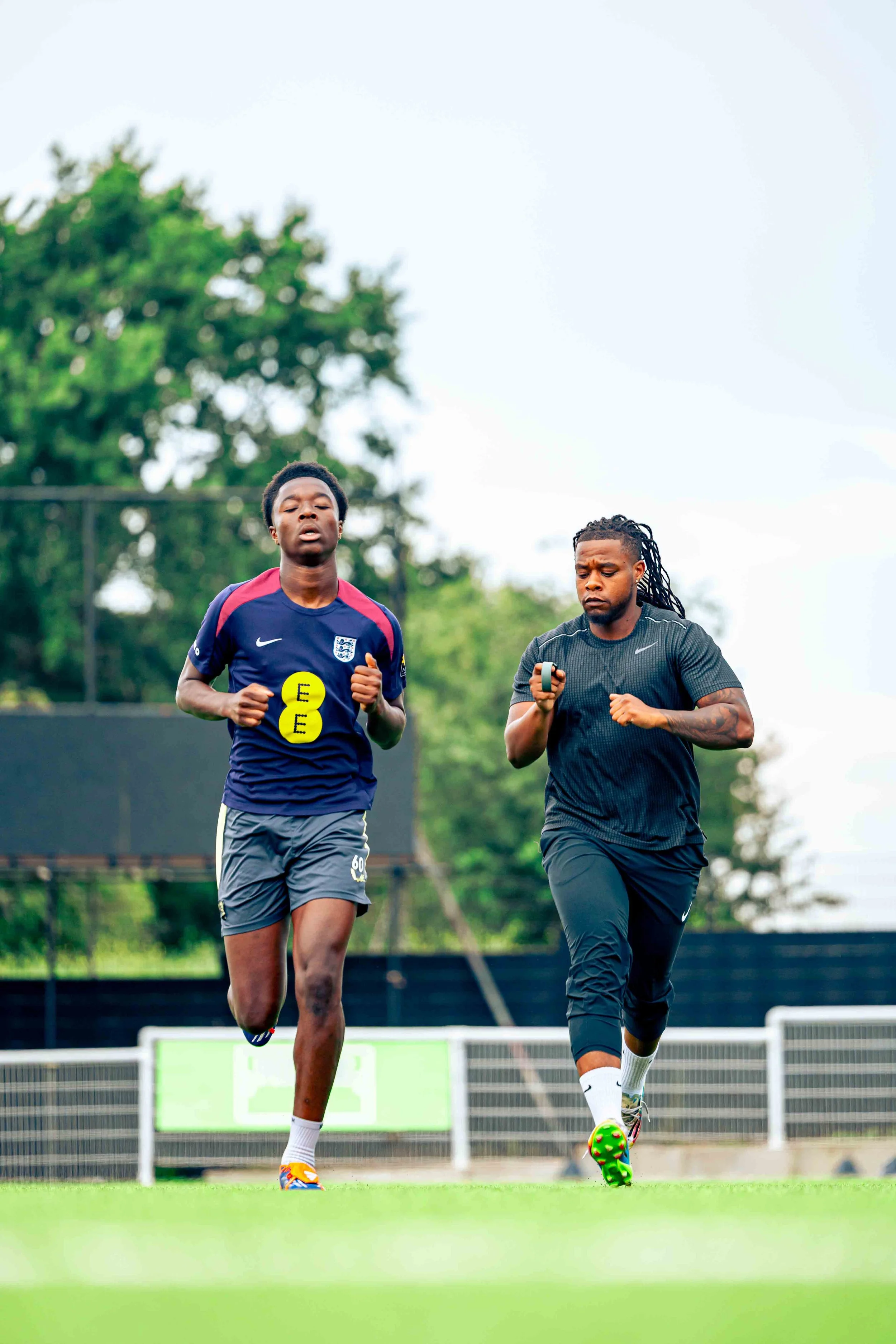 Two male athletes running on a soccer field, with trees and a fence in the background. One athlete is wearing a navy blue jersey with red accents and the England national team logo, while the other is dressed in black athletic wear and holding a devi