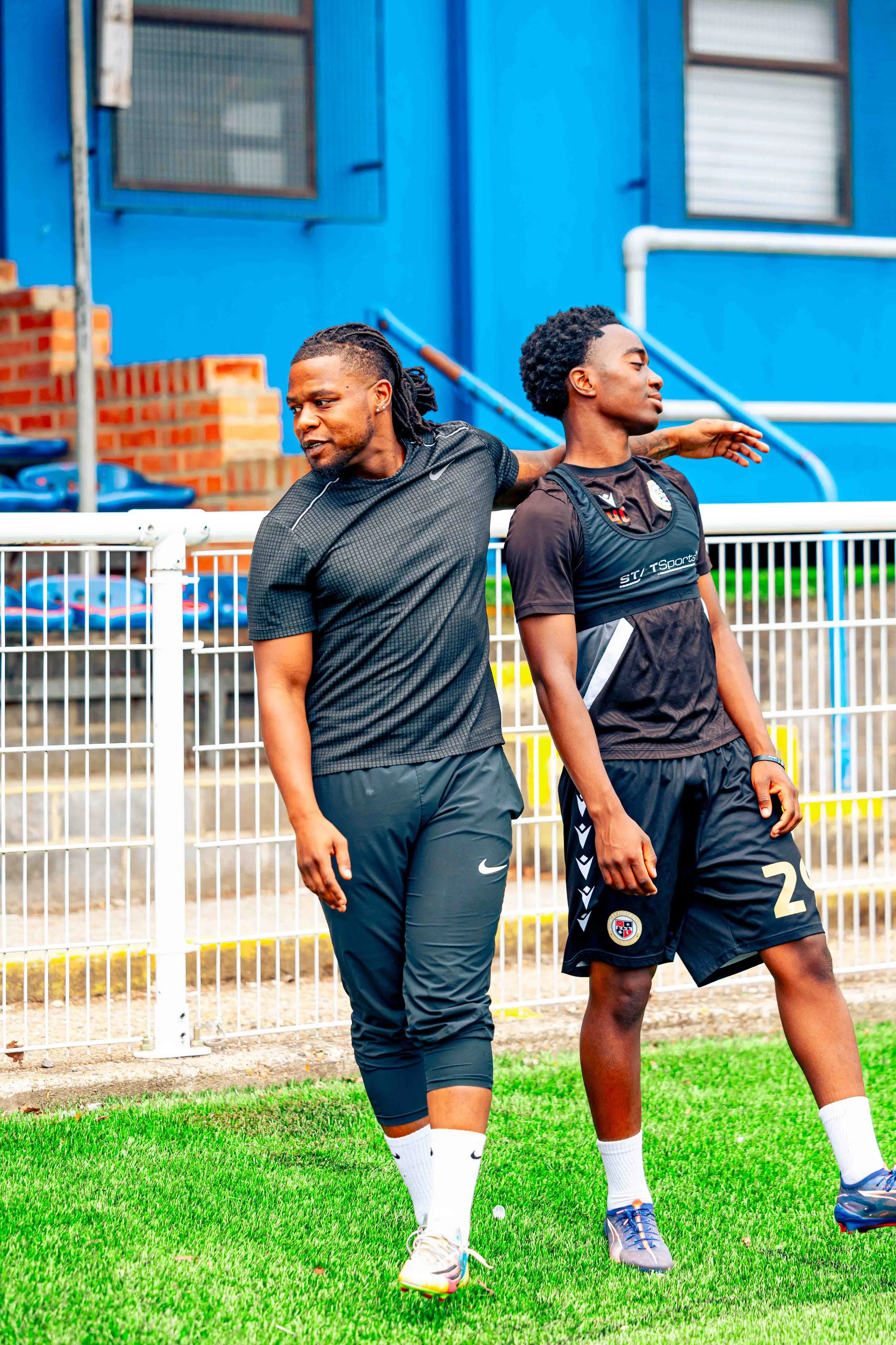 Two soccer players stretching on a soccer field with a blue building and a white fence behind them.