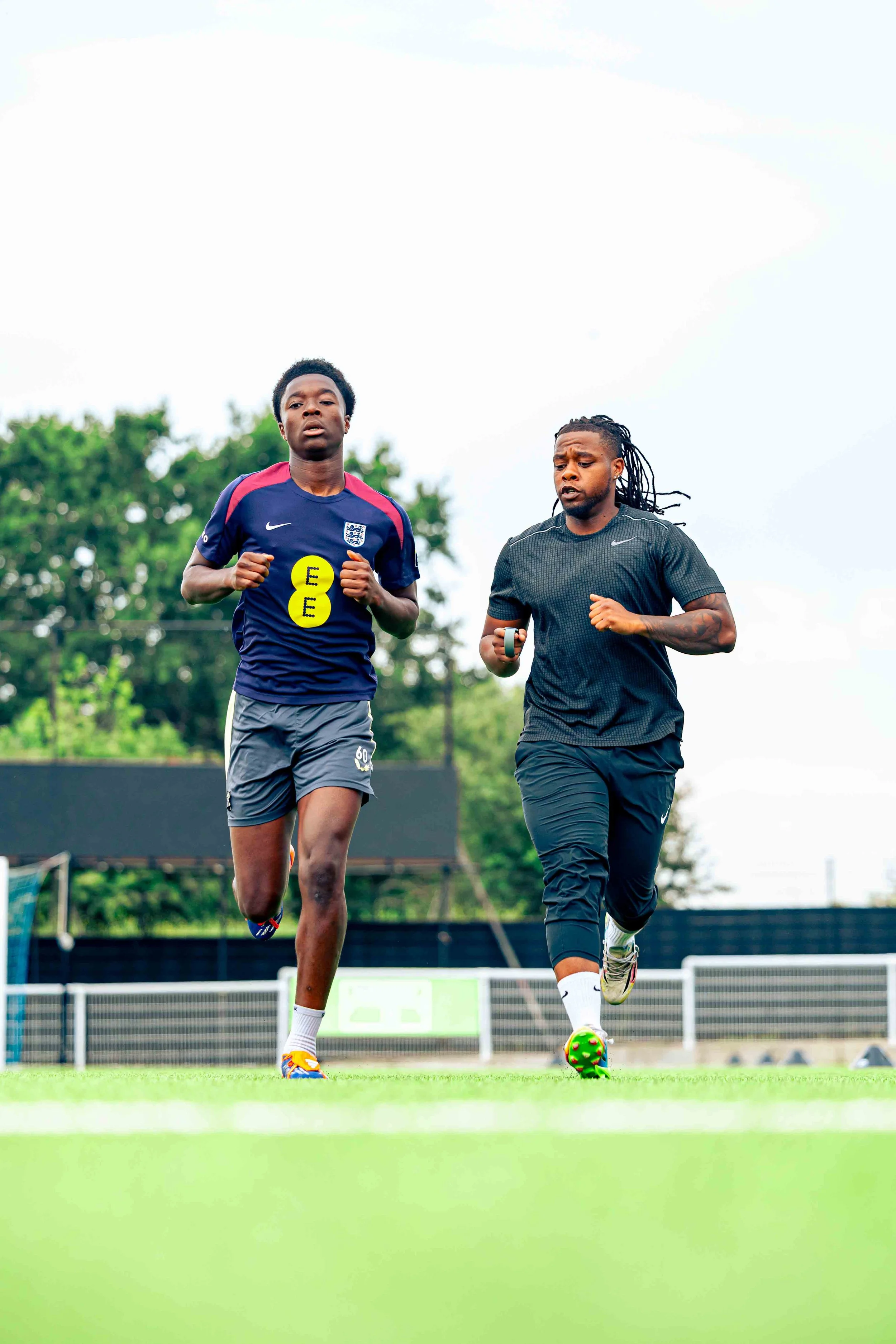 Two men jogging on a green sports field, one wearing a navy blue sports jersey and gray shorts, the other in a black athletic shirt and black pants, with trees in the background and a cloudy sky overhead.