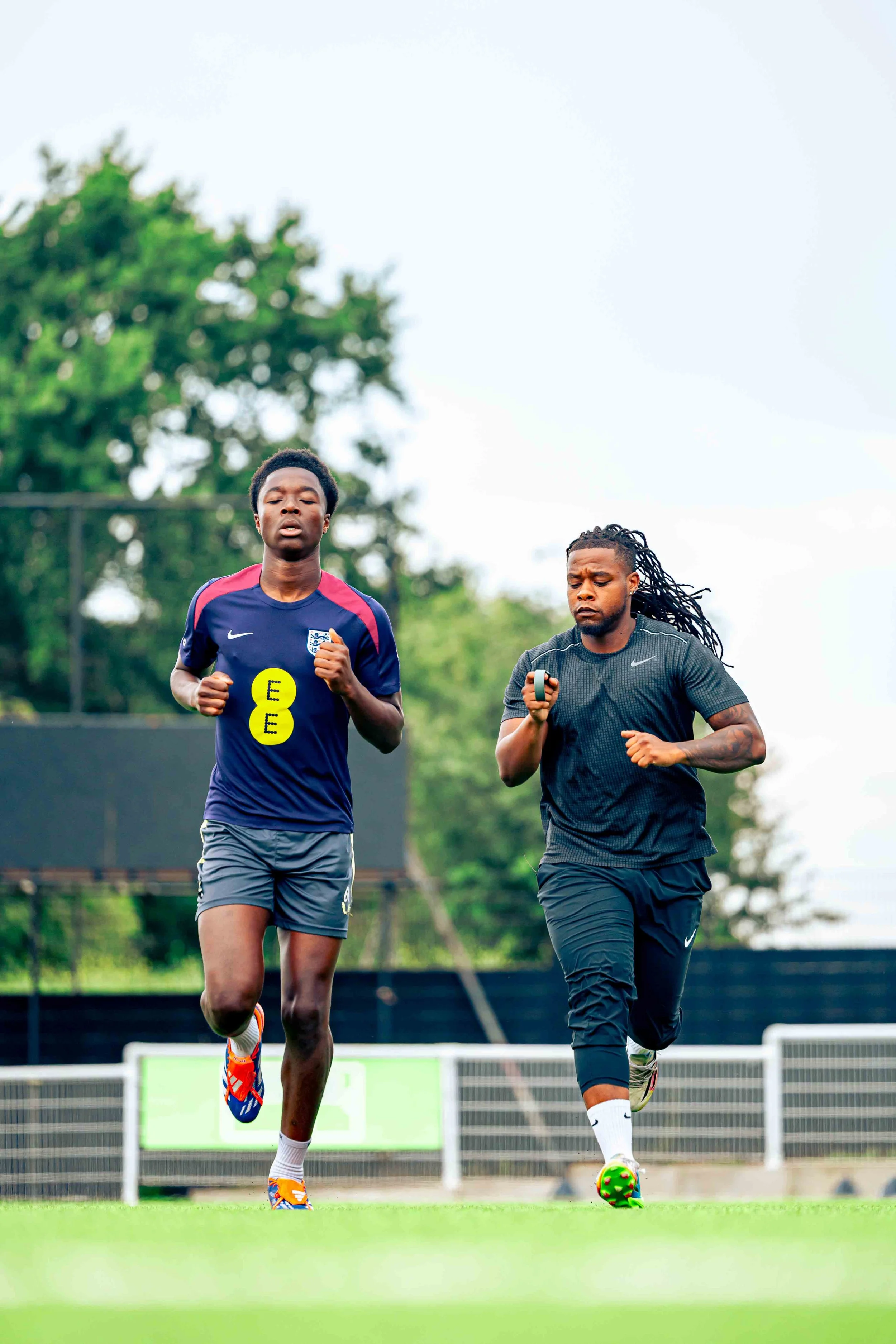 Two men running on a football field during daytime, with trees in the background.
