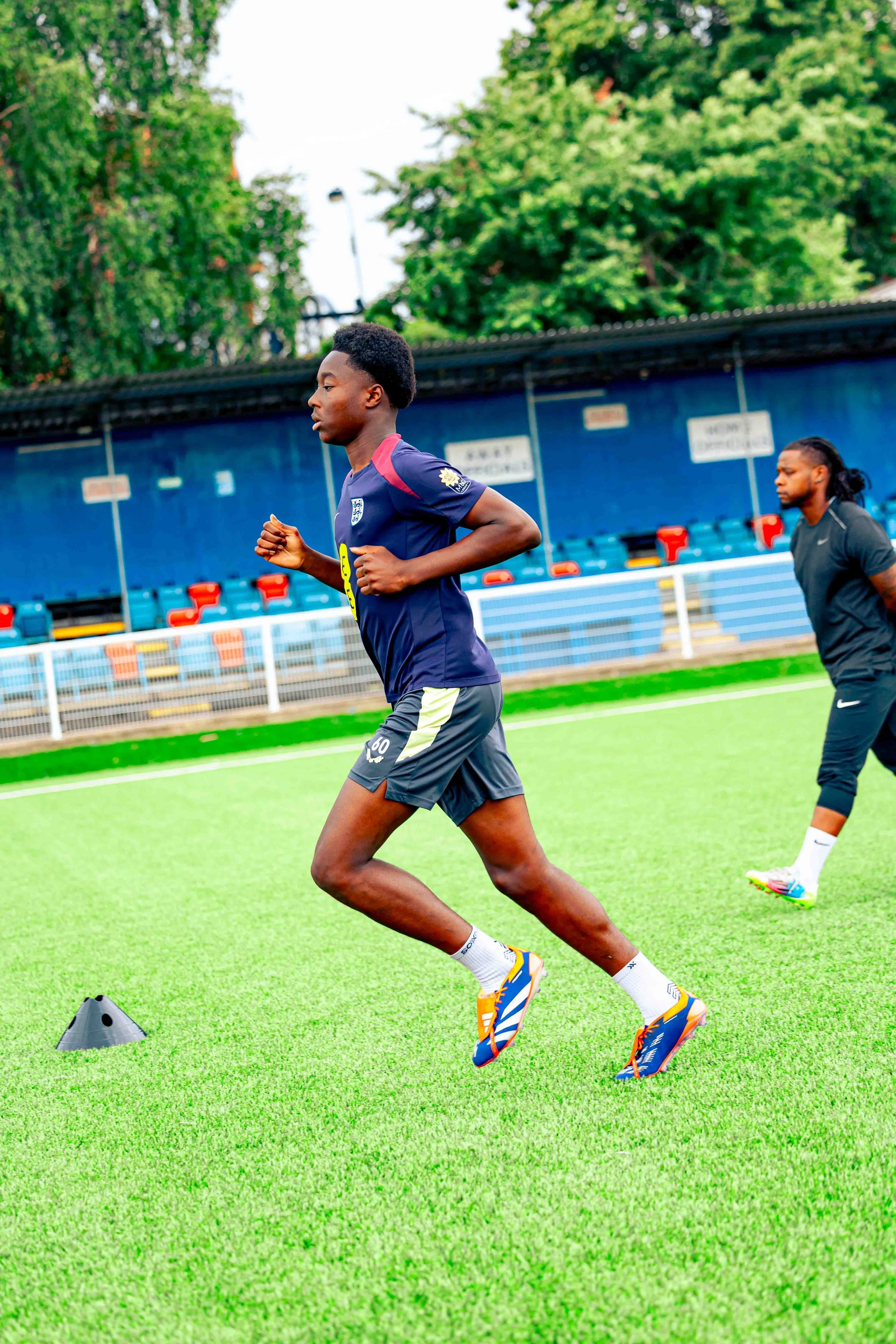 Two young men running on a sports field during a training session, with a background of green trees and a blue stadium seating area.